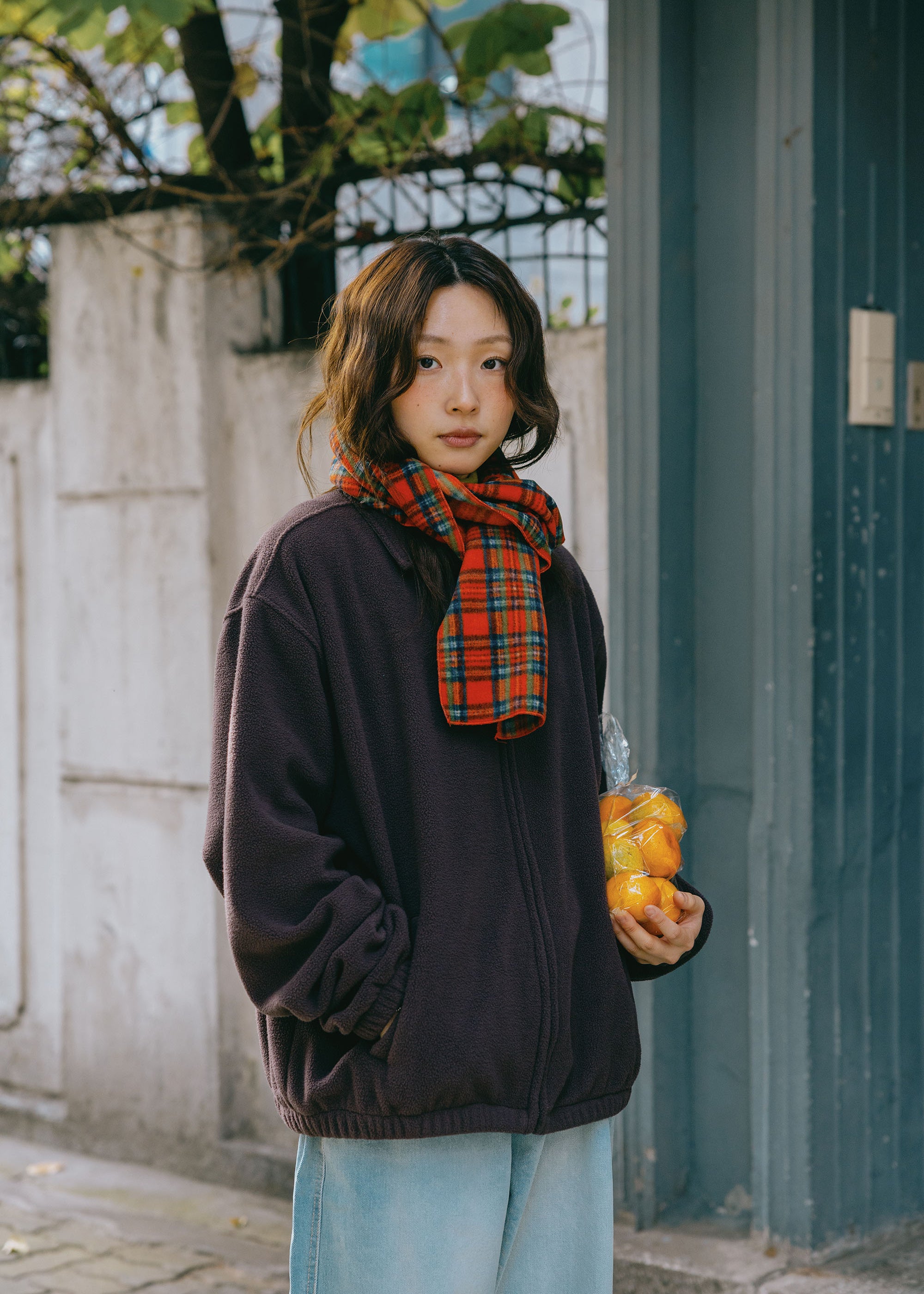 A woman stands outdoors wearing a dark brownish-purple zip-up jacket and a red and black plaid scarf, holding a clear bag of oranges. She is positioned in front of a gray concrete wall and a dark blue-gray wooden door.