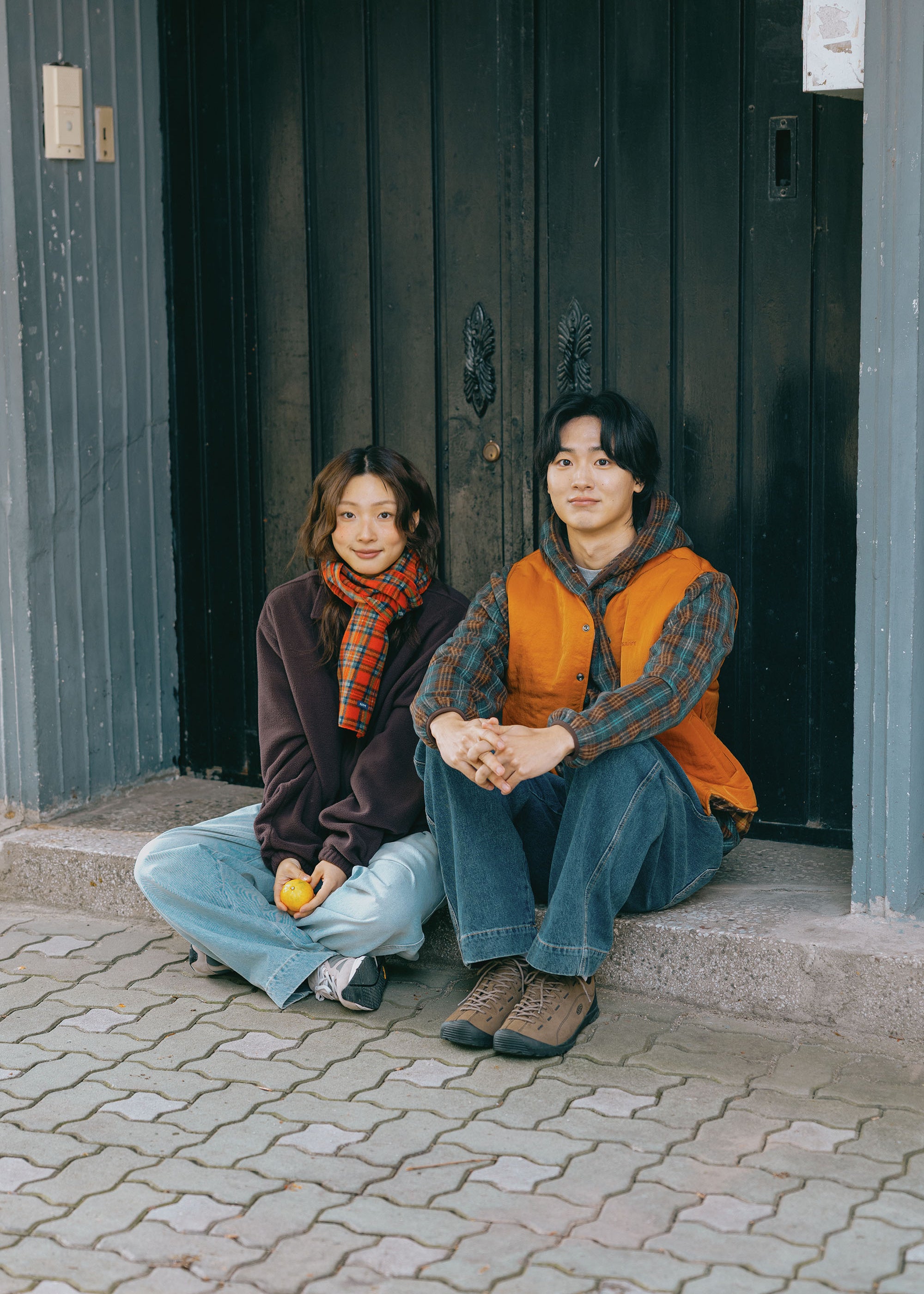 Two young people sitting on stone steps in front of a dark wooden door. The person on the left wears a dark brown hoodie, a red and black plaid scarf, and light blue jeans.