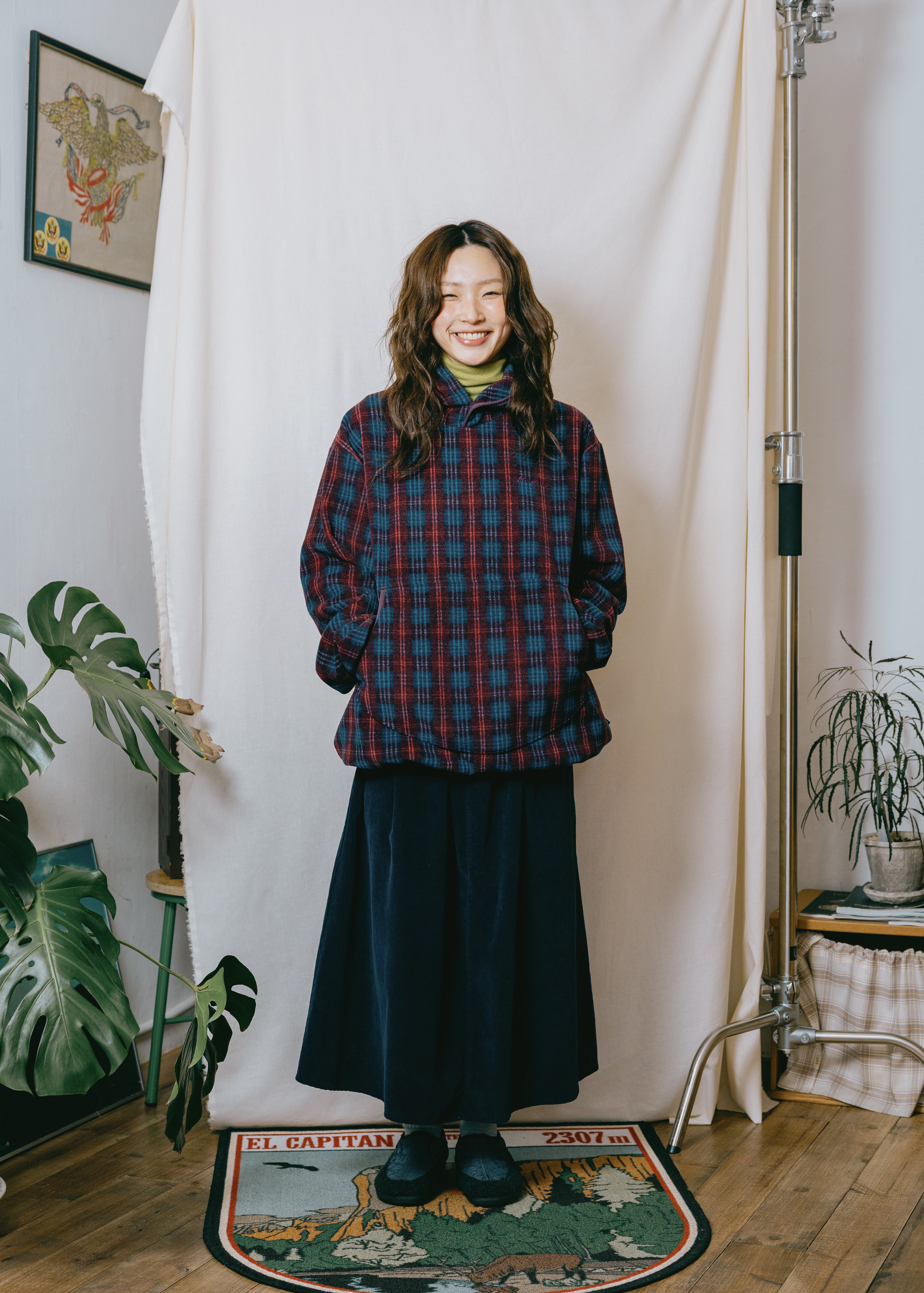 A woman stands in front of a white fabric backdrop in a studio-like room, wearing a red and navy plaid overshirt over a yellow top and a long dark skirt. The scene includes plants, a framed print on the wall, and a patterned rug on a wooden floor.