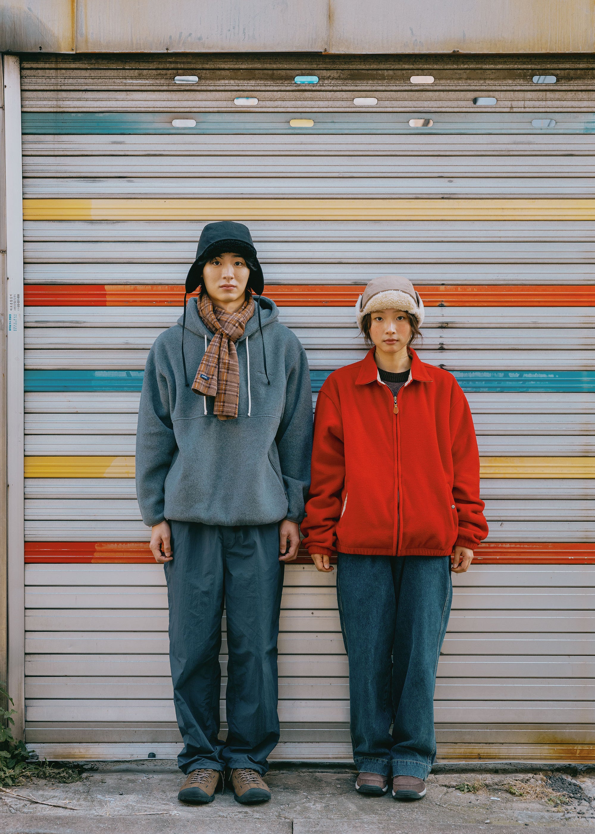 Two people standing side by side in front of a closed metal roll-up garage door with horizontal stripes in yellow, orange, blue, and red. The person on the left wears a dark trapper hat, a gray hoodie, a brown scarf, and dark pants.