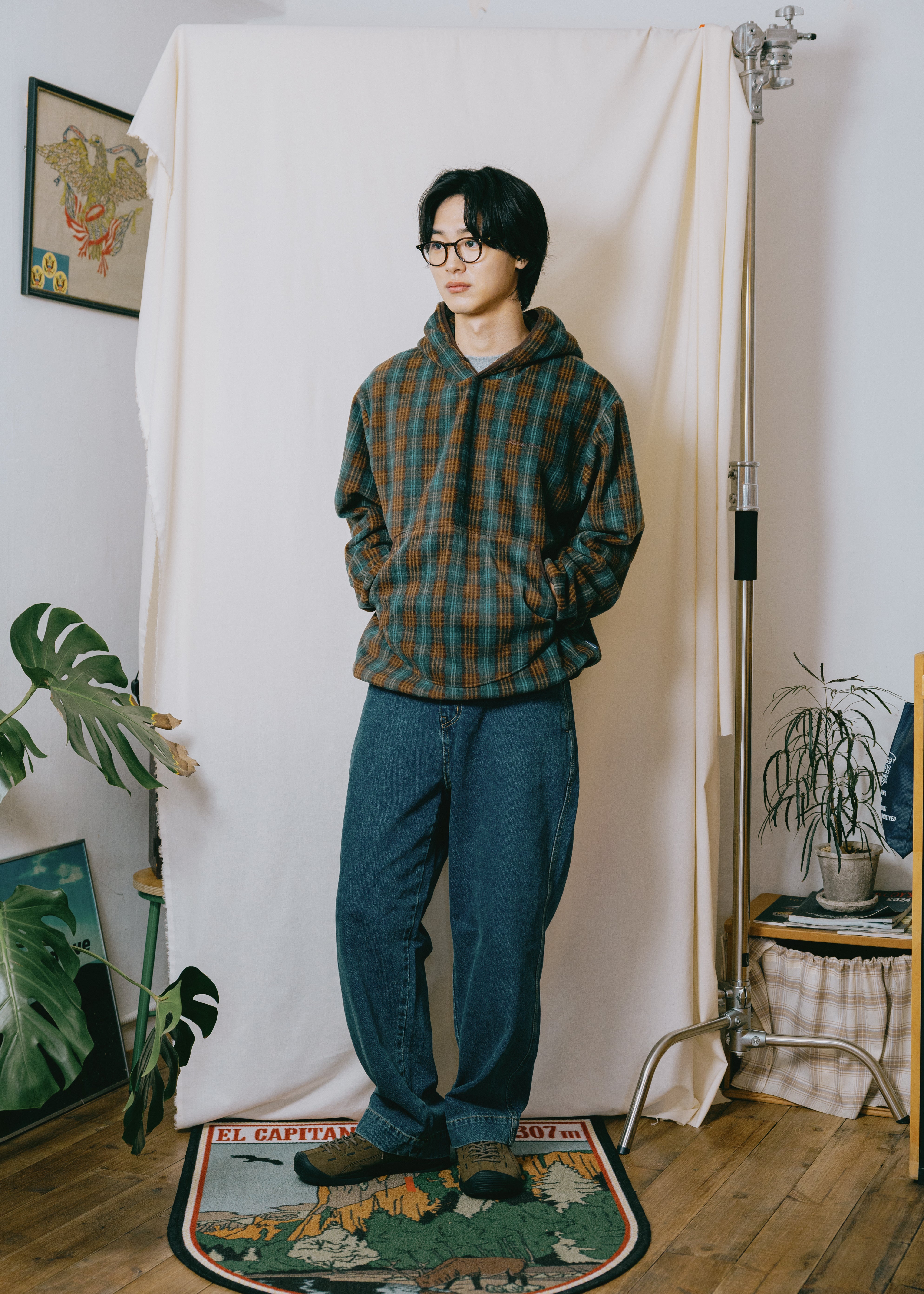 A man wearing a brown and green plaid hoodie, dark blue jeans, and glasses stands in a studio setting with a white fabric backdrop, wooden floor, and plants.