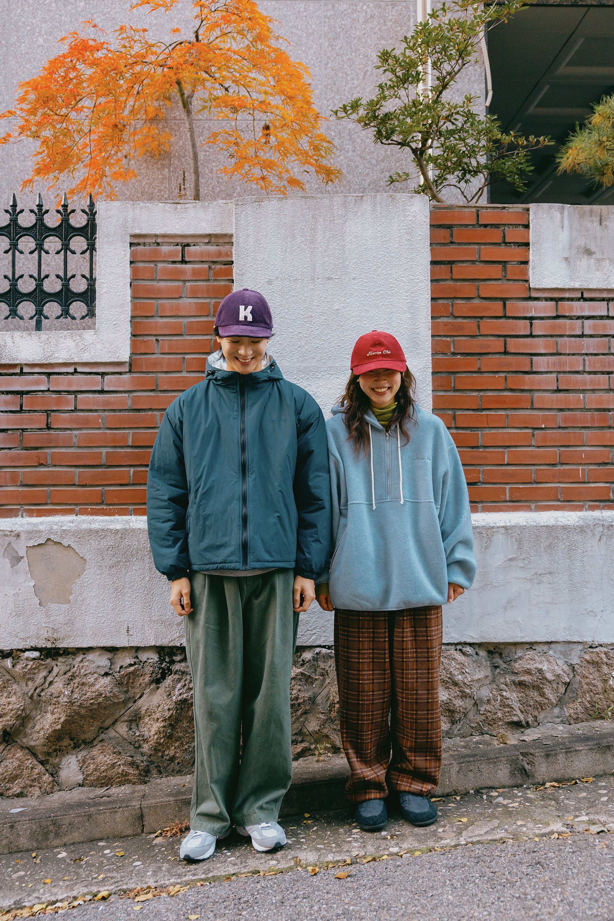 Two people standing outdoors in front of a brick wall and stone foundation, wearing casual streetwear. The person on the left wears a dark teal zip-up jacket, olive green pants, and a purple cap with a white letter 'K'. the woman on the right is a light blue fleece hoodie