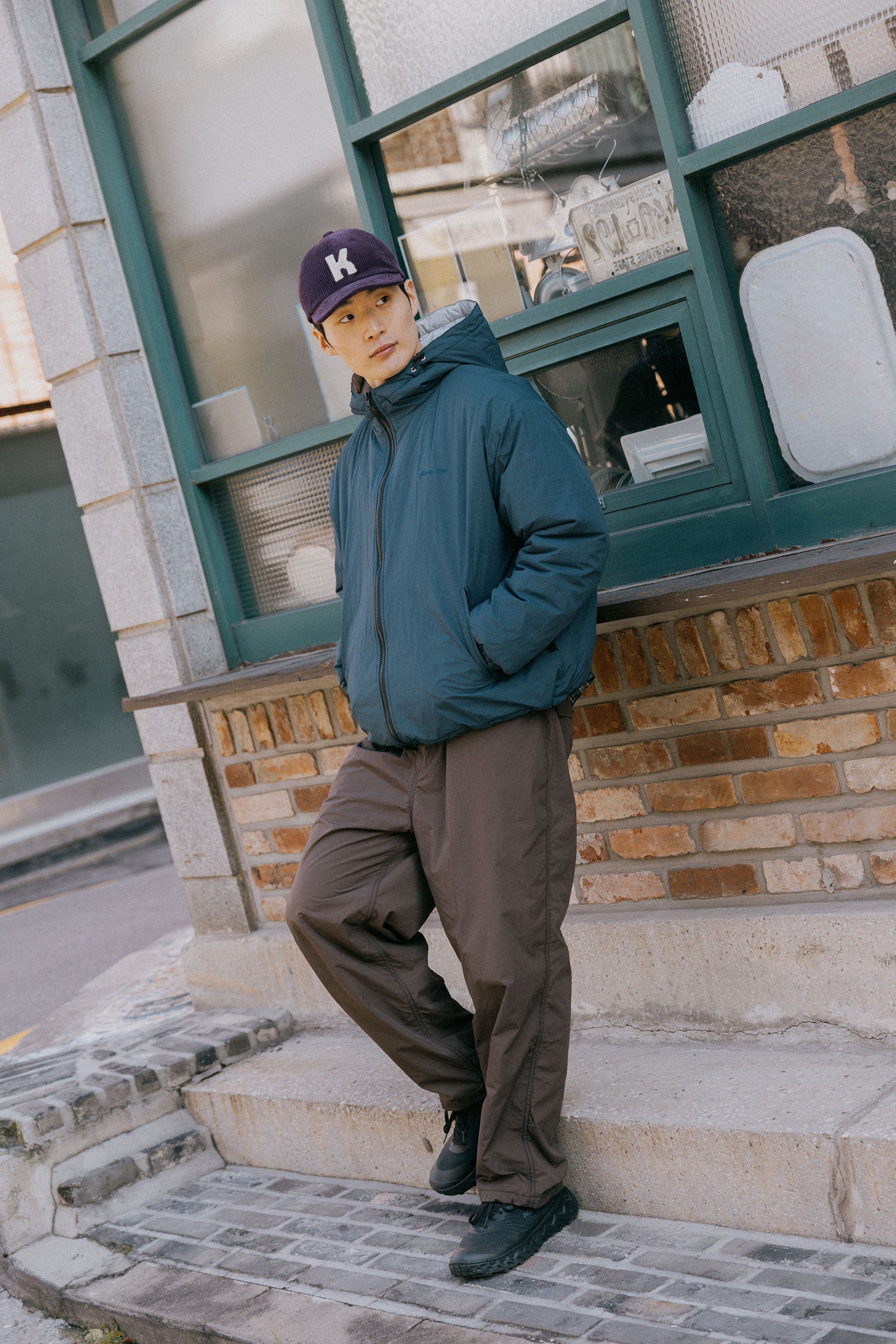 A man wearing a dark teal puffer jacket, brown pants, and a purple baseball cap leans against the stone steps of a building with green-framed windows.