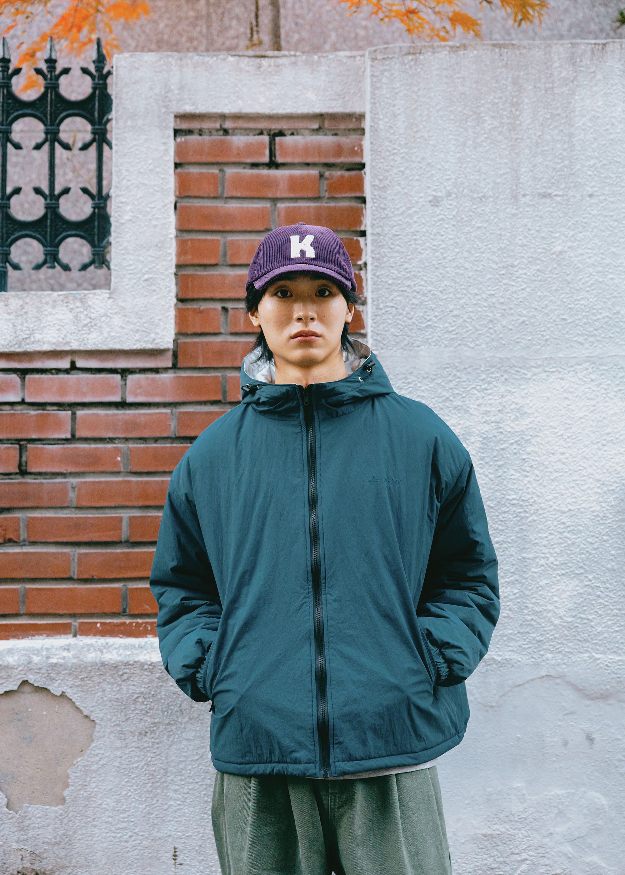 A person standing outdoors in front of a white and red brick wall, wearing a dark teal zip-up puffer jacket, olive green pants, and a purple baseball cap with a white letter 'K'.