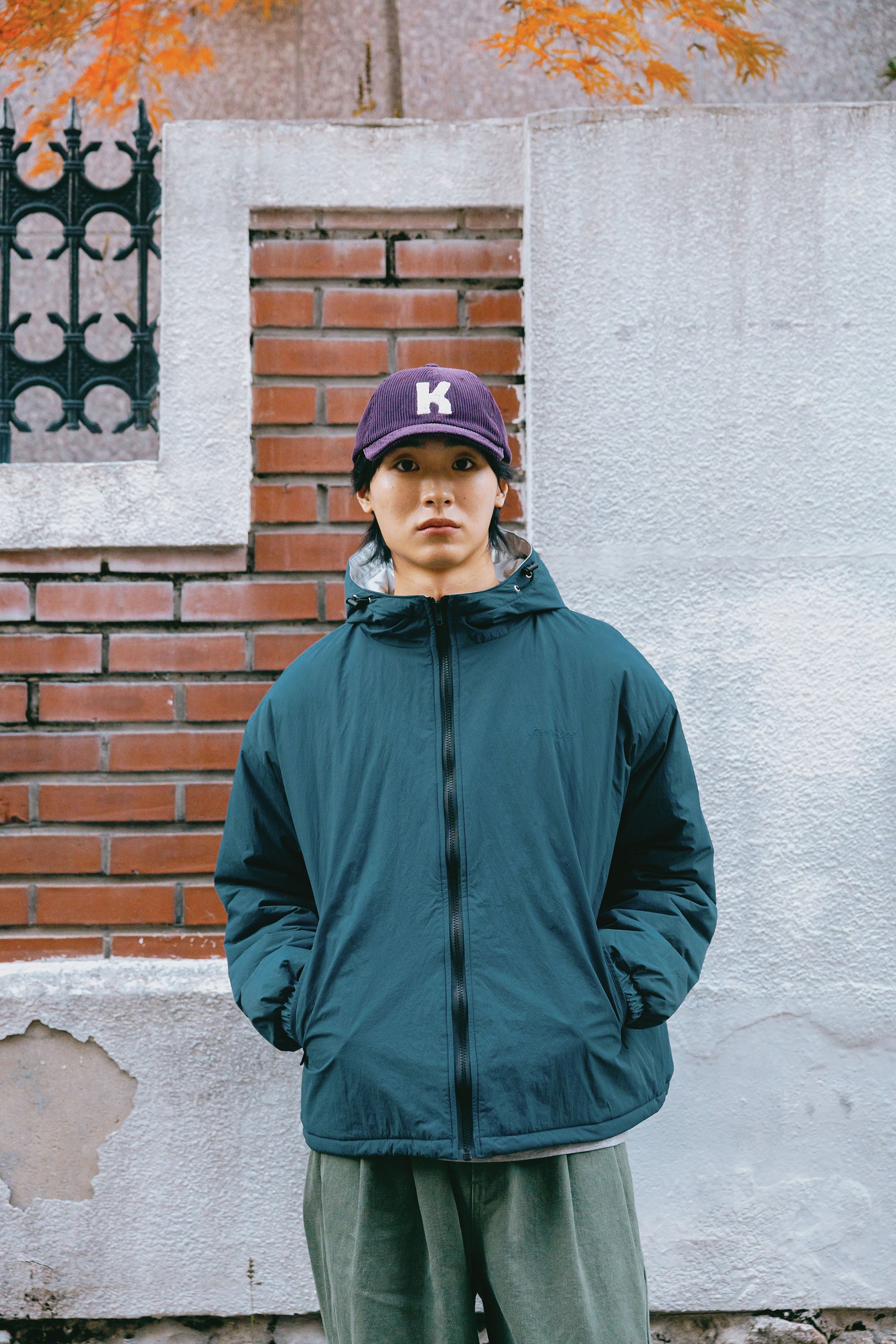 A person standing outdoors in front of a white and red brick wall, wearing a dark teal zip-up puffer jacket, olive green pants, and a purple baseball cap with a white letter 'K'.