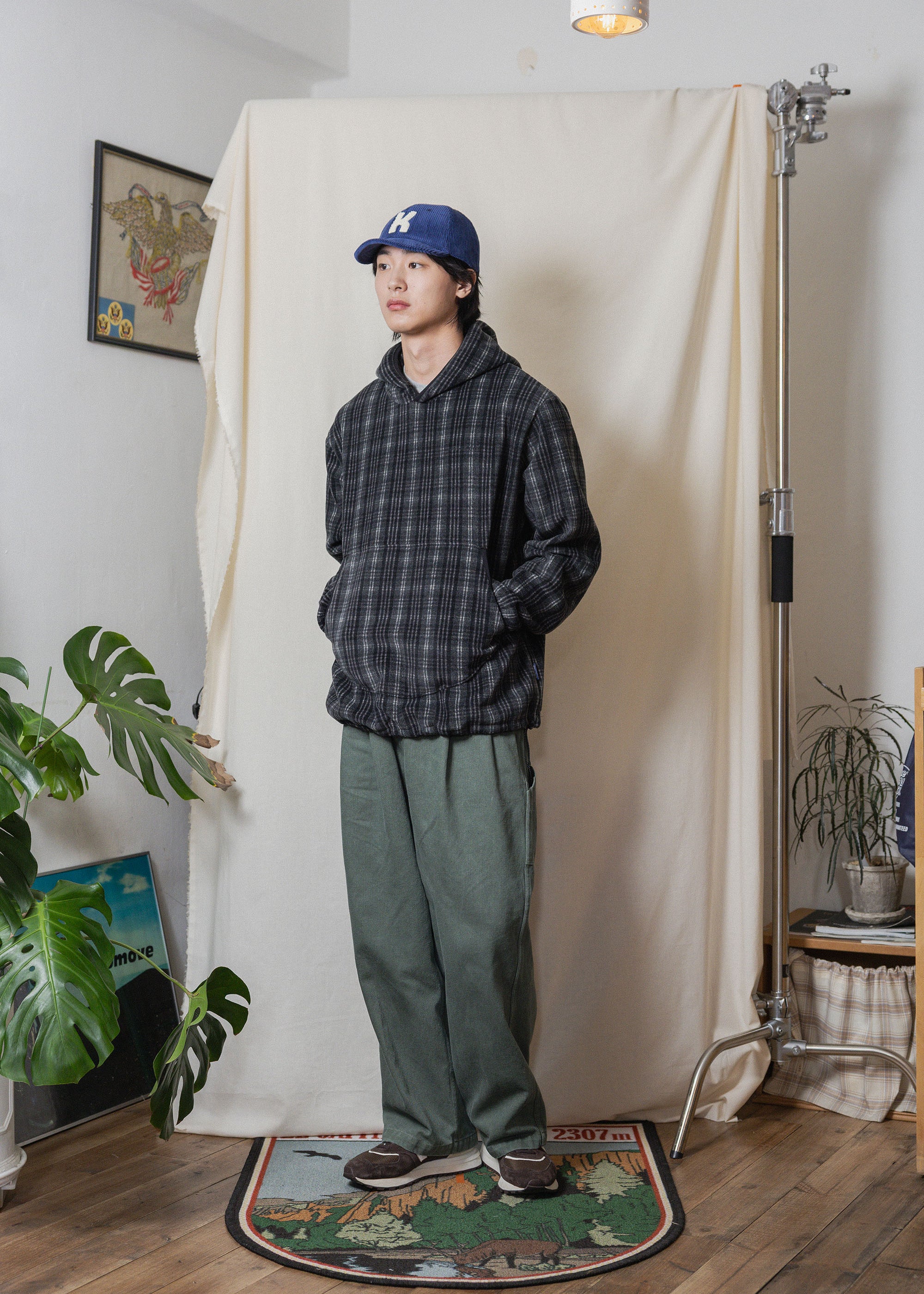 A person stands in a studio setup wearing a dark plaid hooded jacket, olive green pants, and a blue baseball cap, positioned in front of a cream fabric backdrop with plants and floor lamps visible around the room.
