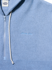 Close-up of a light blue heathered quarter-zip hoodie with a white drawstring and subtle tonal embroidery on the chest. A white brand label is visible at the collar.