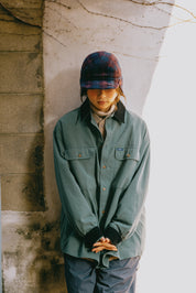 A person wearing a muted sage green button-up overshirt, dark plaid cap, and gray pants, standing against a textured concrete wall in natural light.