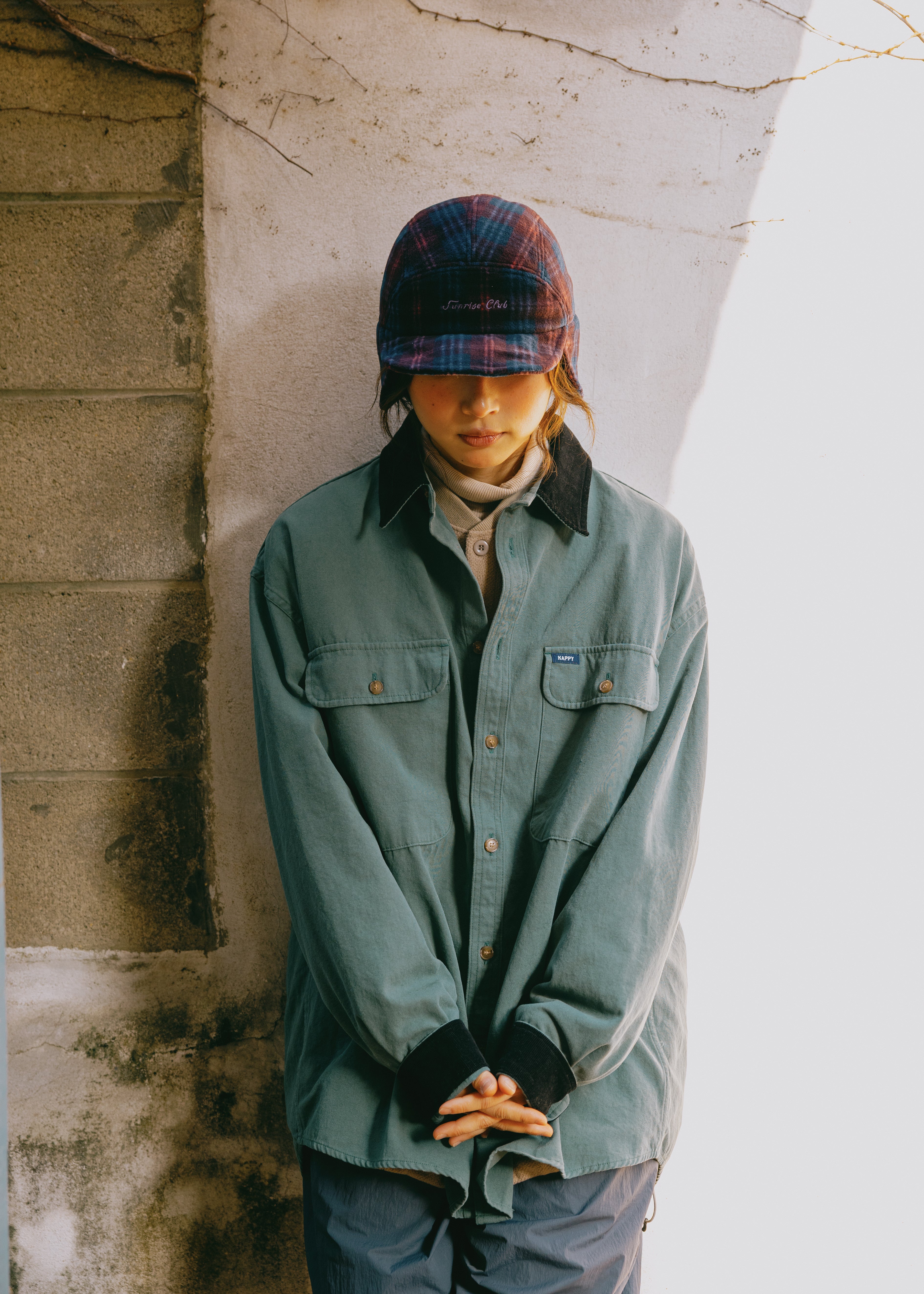 A person wearing a muted sage green button-up overshirt, dark plaid cap, and gray pants, standing against a textured concrete wall in natural light.