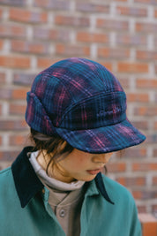 a woman wearing plaid 5-star cap with ear flaps in a dark blue and purple pattern, featuring white embroidered text on the front that reads 'Sunrise Club'.