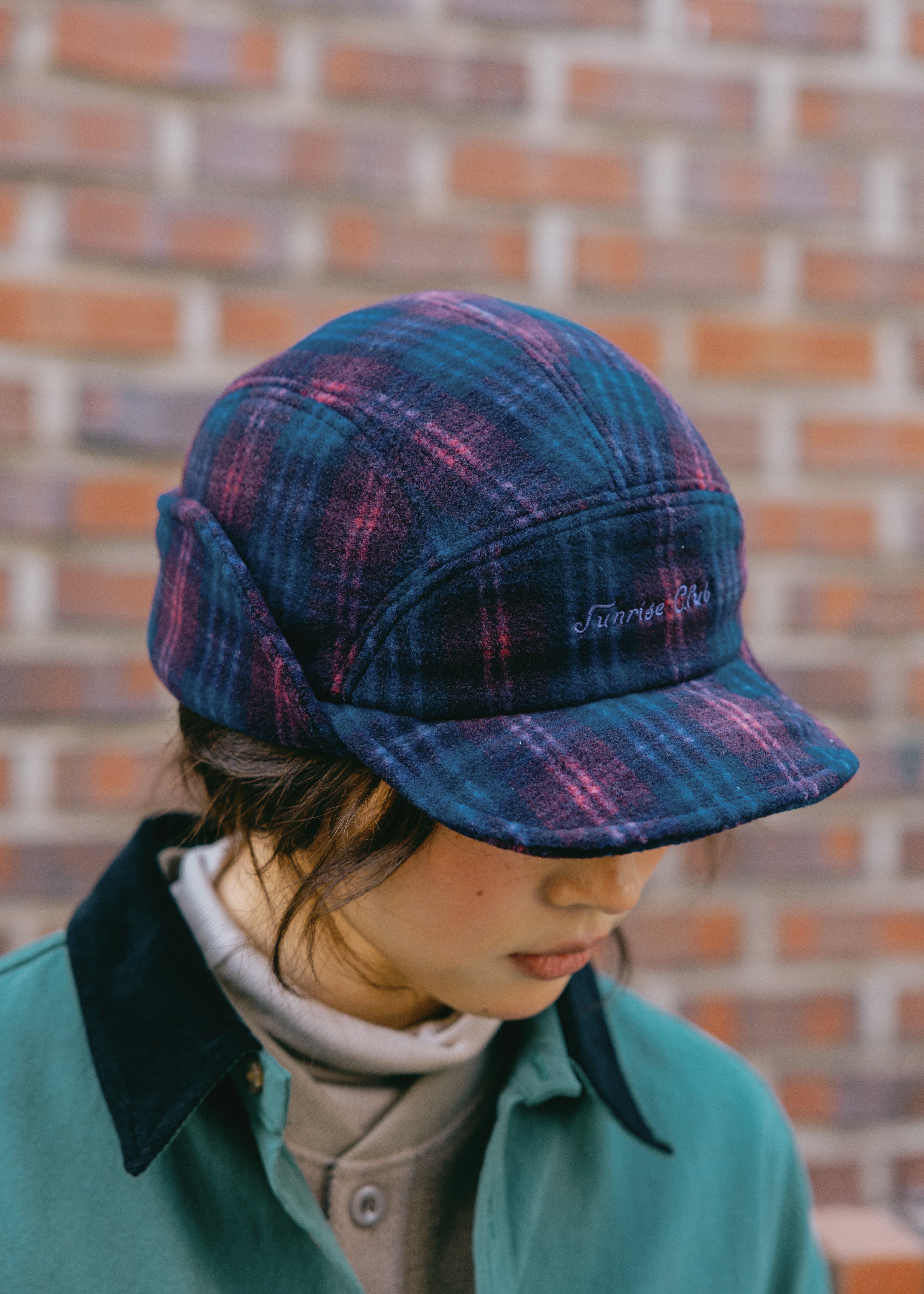 a woman wearing plaid 5-star cap with ear flaps in a dark blue and purple pattern, featuring white embroidered text on the front that reads 'Sunrise Club'.