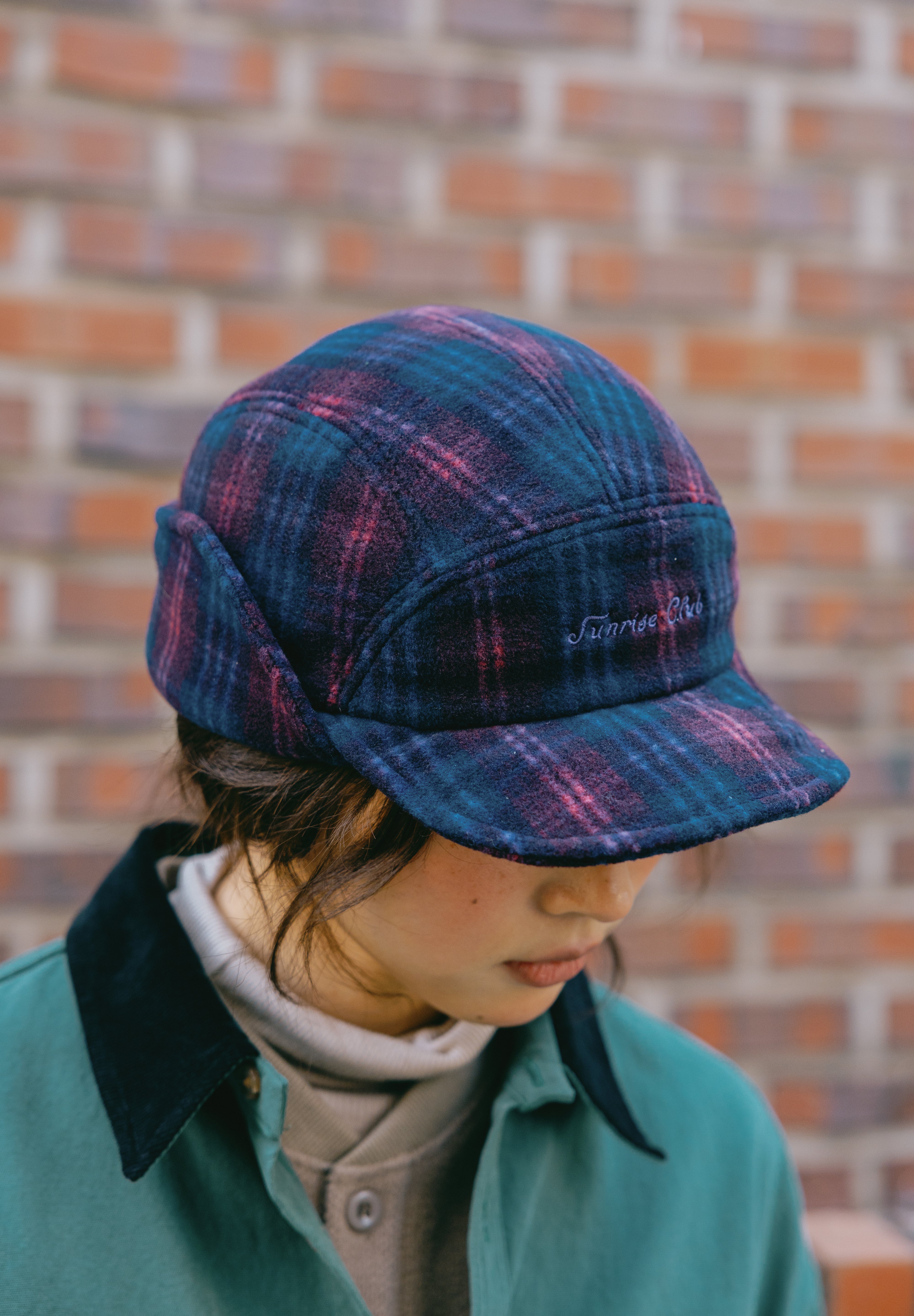 a woman wearing plaid 5-star cap with ear flaps in a dark blue and purple pattern, featuring white embroidered text on the front that reads 'Sunrise Club'.