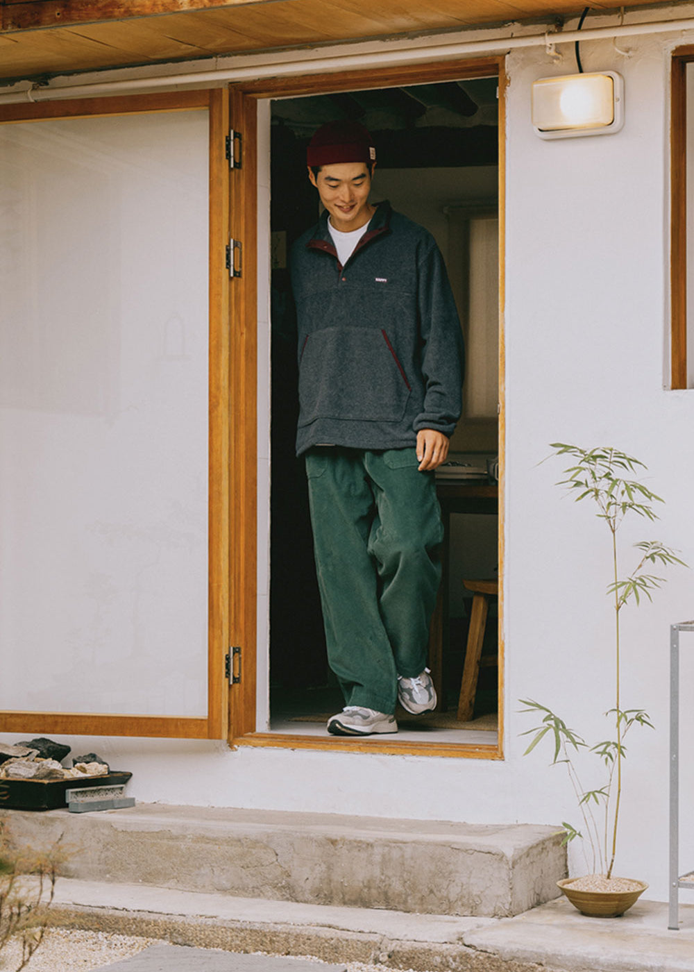 Person standing in a doorway wearing a dark green fleece and green pants, with a plant and light fixture visible.