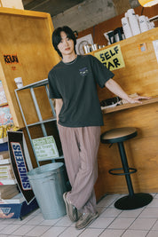 Person wearing a dark green t-shirt and light-colored pants in a casual setting.