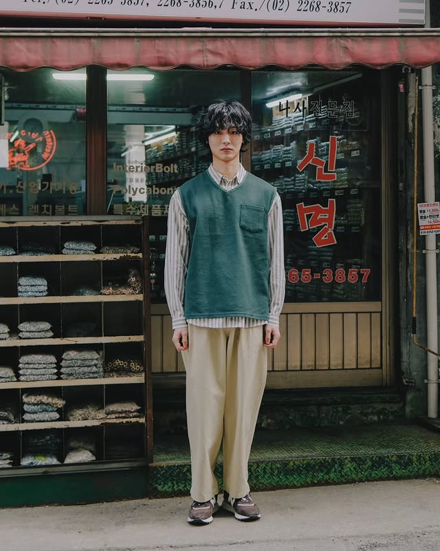Person wearing a green vest and beige pants standing in front of a store with Korean text.