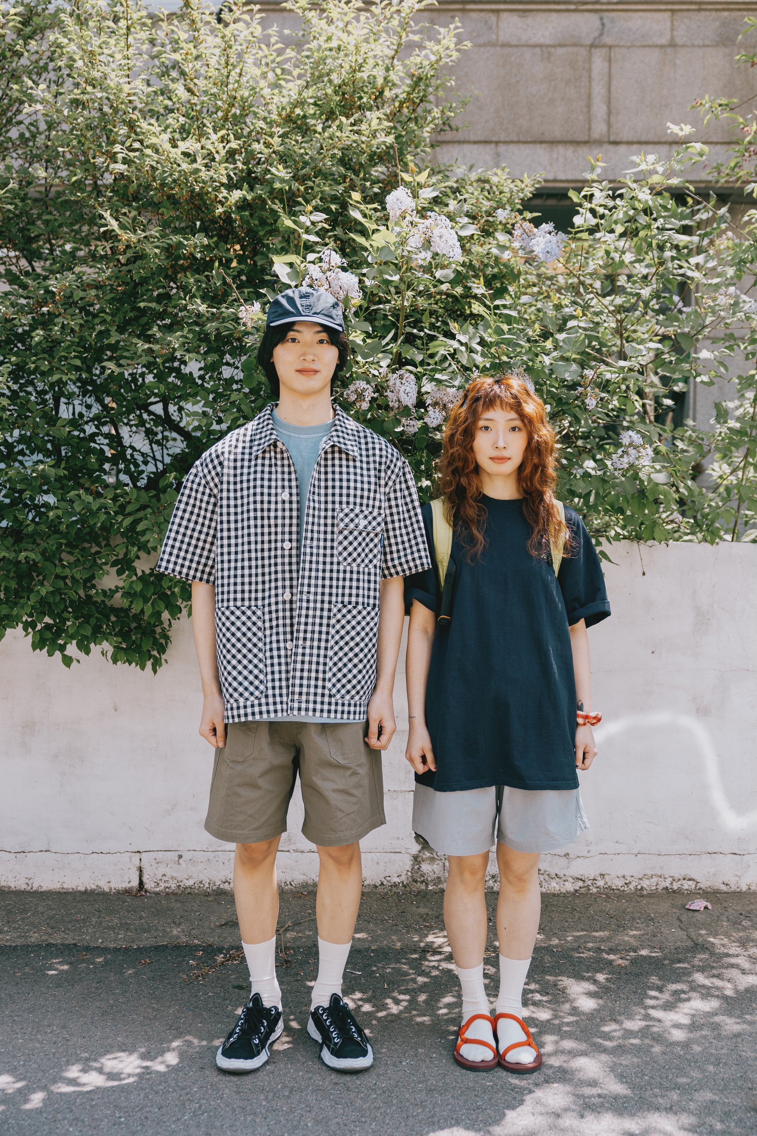 Two people standing outdoors with greenery and a building in the background