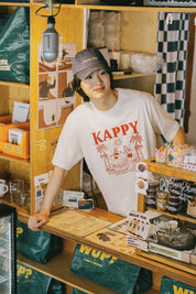 Person wearing a 'Kappy' t-shirt in a store setting with various items on shelves.