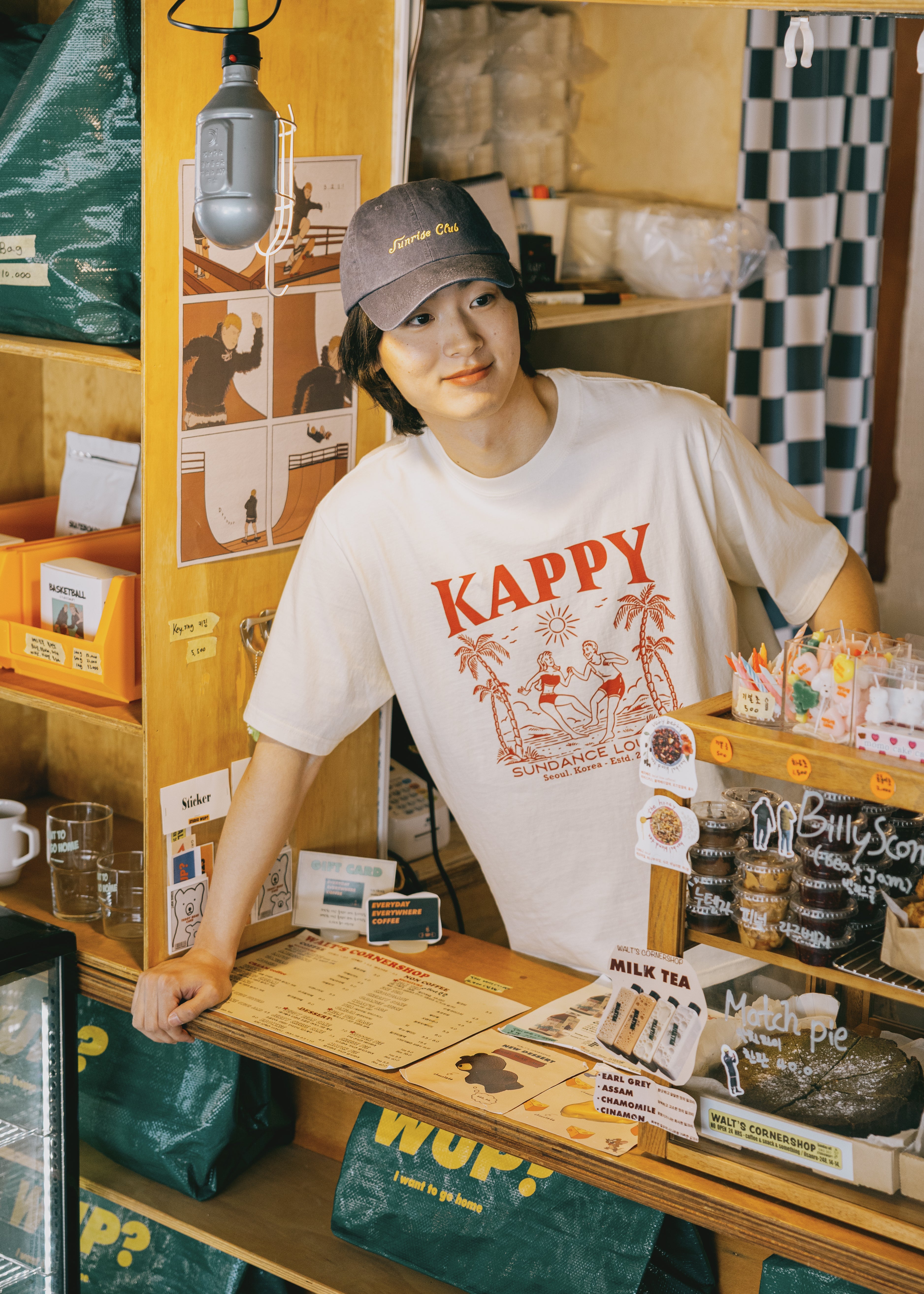 Person wearing a 'Kappy' t-shirt in a store setting with various items on shelves.
