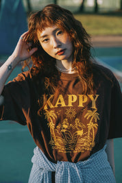 Person wearing a brown t-shirt with 'Kappy' design, standing by basketball court.