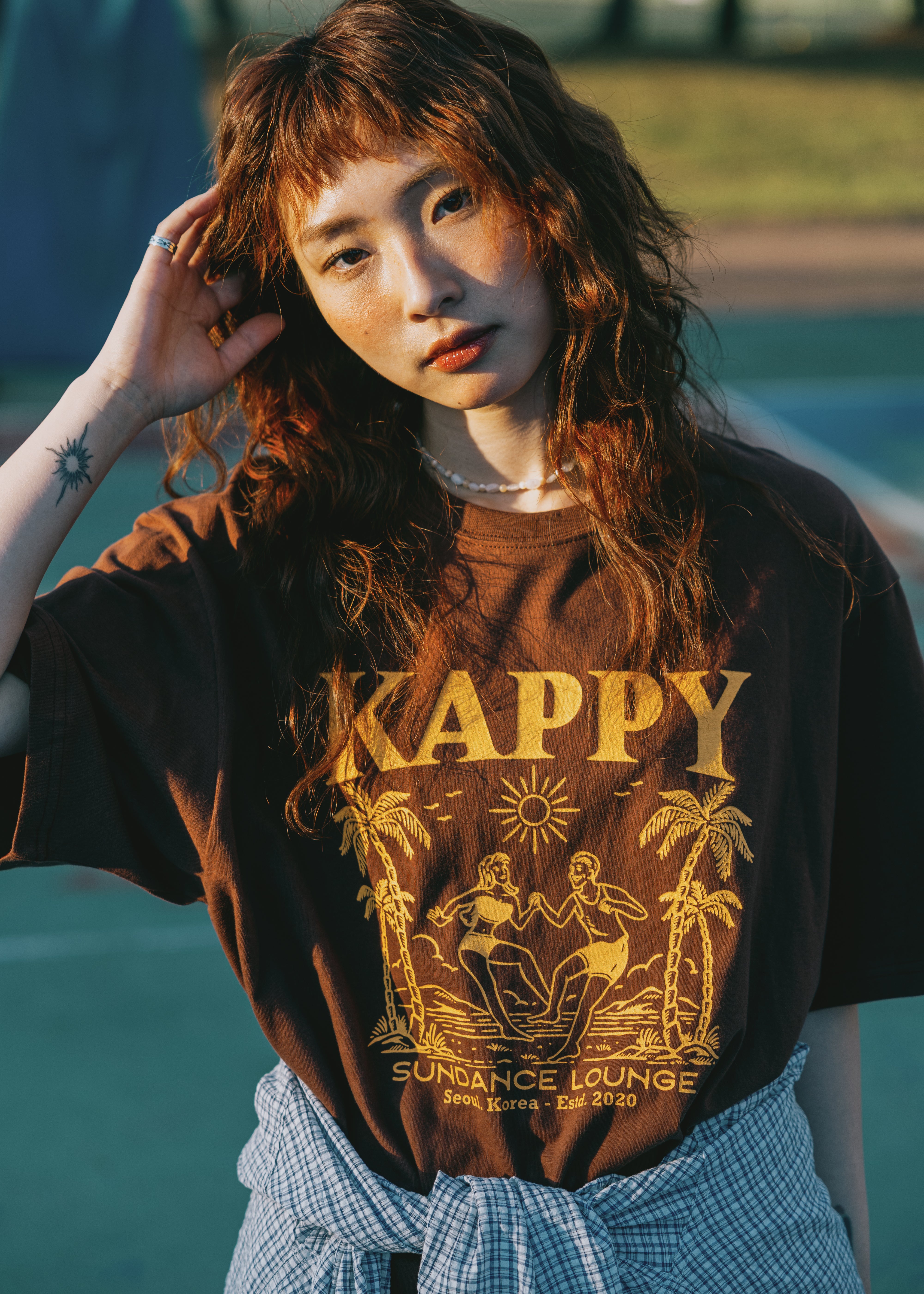 Person wearing a brown t-shirt with 'Kappy' design, standing by basketball court.