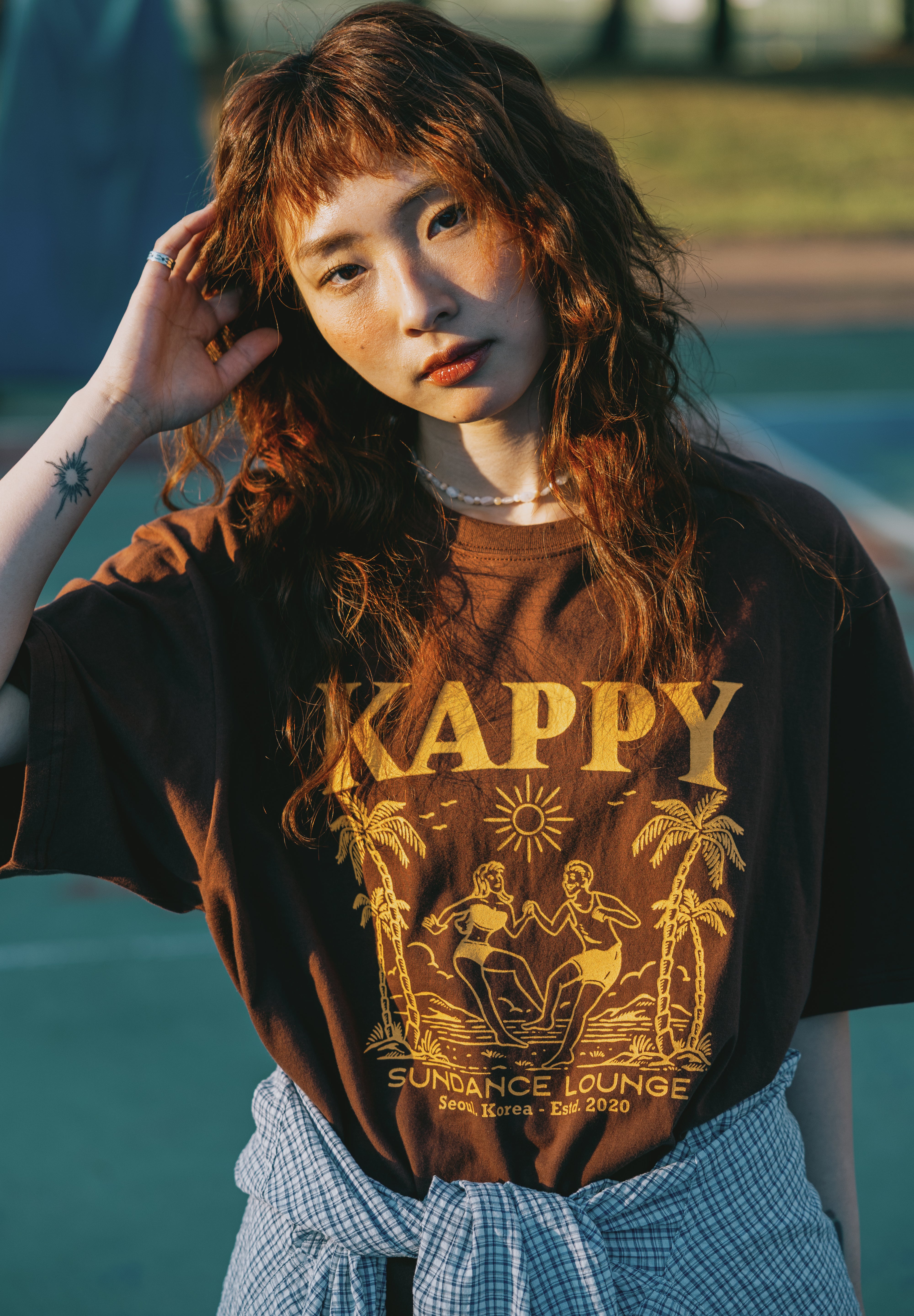 Person wearing a brown t-shirt with 'Kappy' design, standing by basketball court.