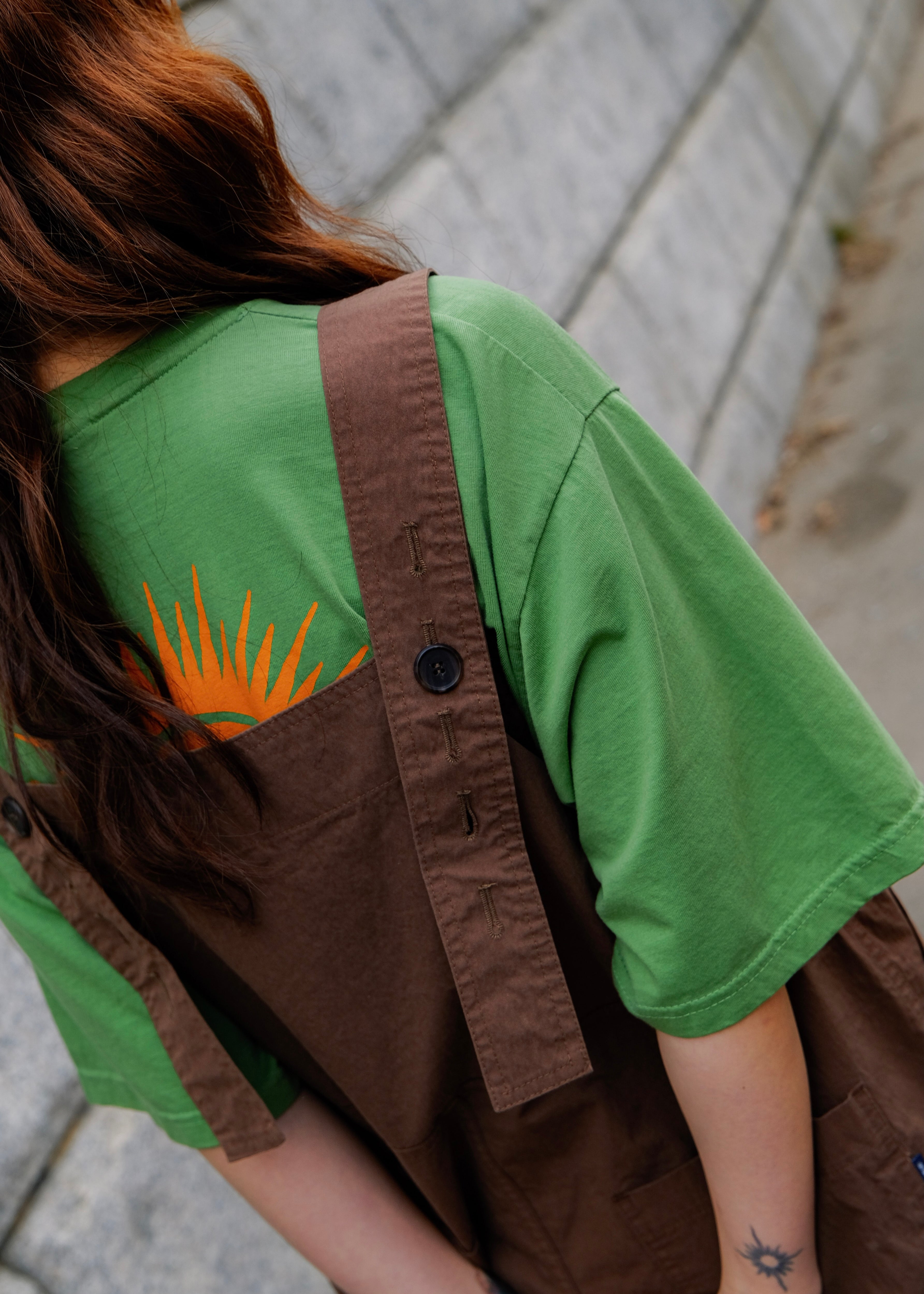 Person wearing a green shirt with a rays of the sun print and brown overalls sitting on a stone bench.