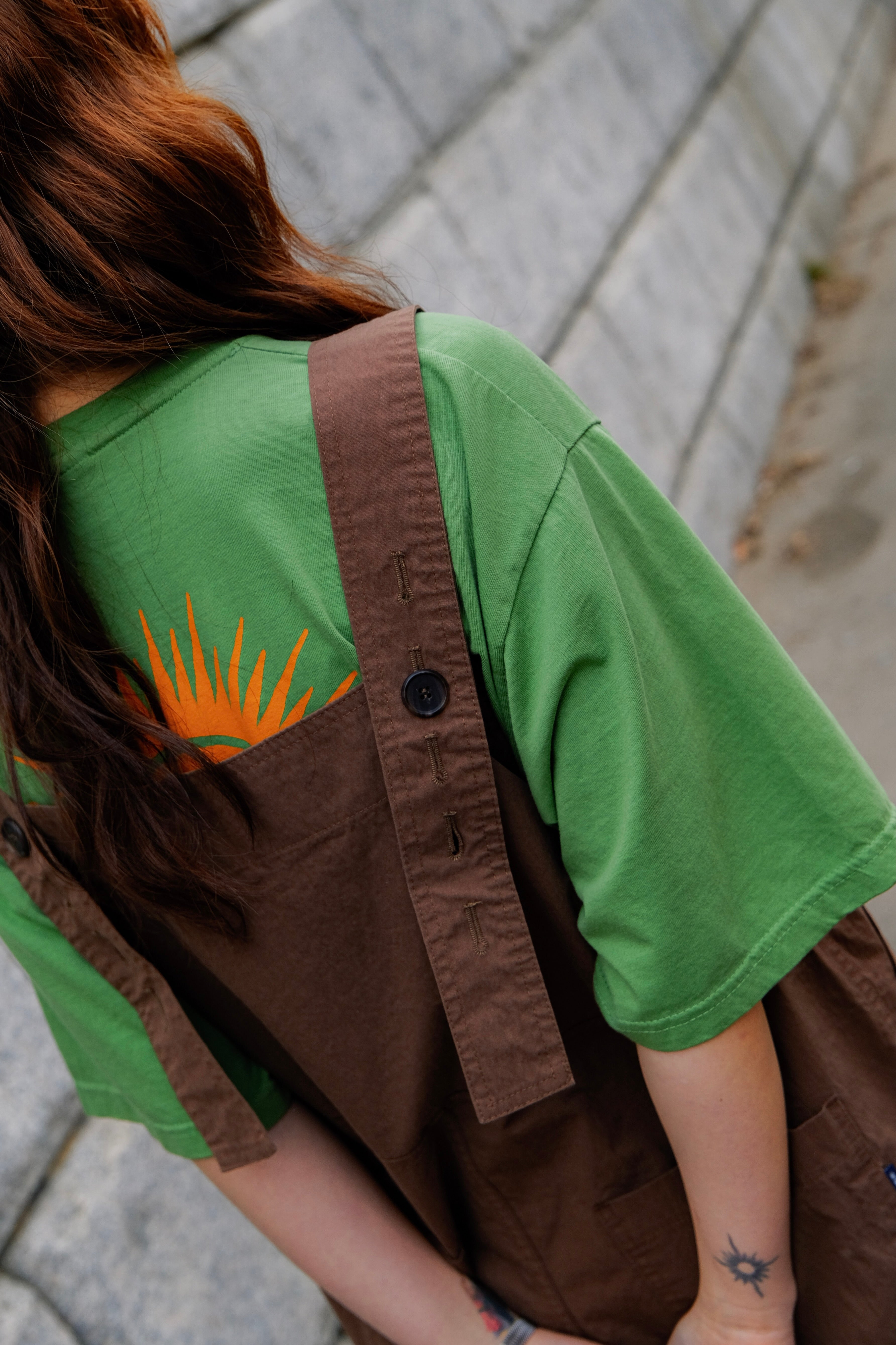 Person wearing a green shirt with a rays of the sun print and brown overalls sitting on a stone bench.