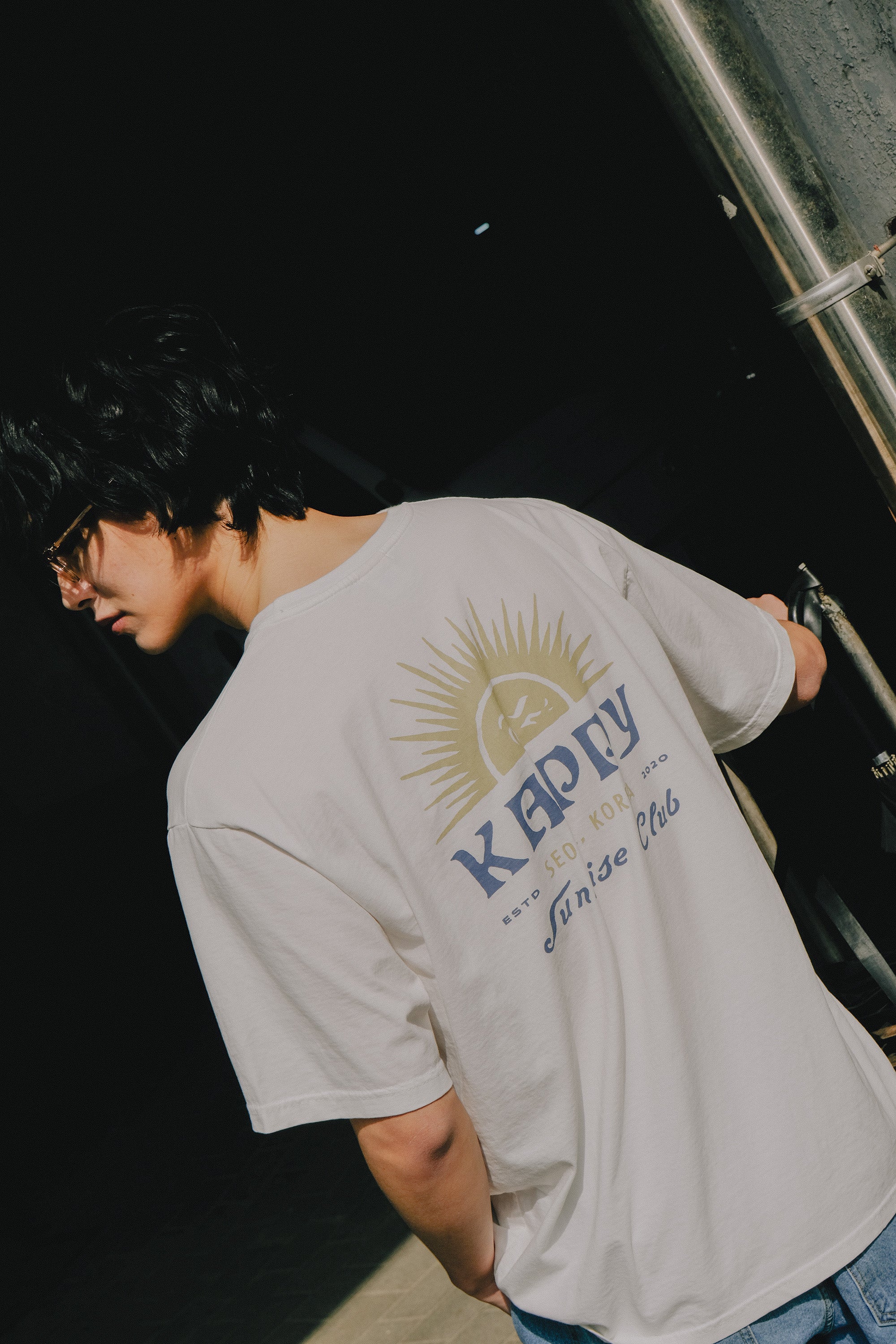 Person wearing a white t-shirt with a logo and text, standing against a dark background.