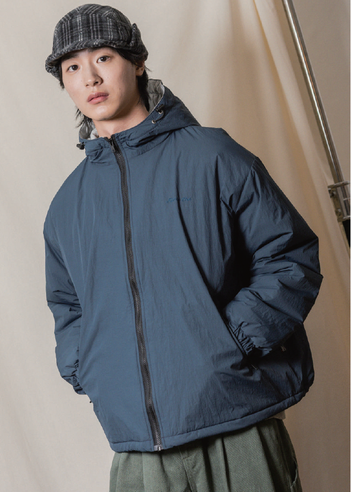 A model wearing a dark slate blue hooded puffer jacket, a dark plaid fleece hat, and olive green pants, standing against a neutral beige backdrop.