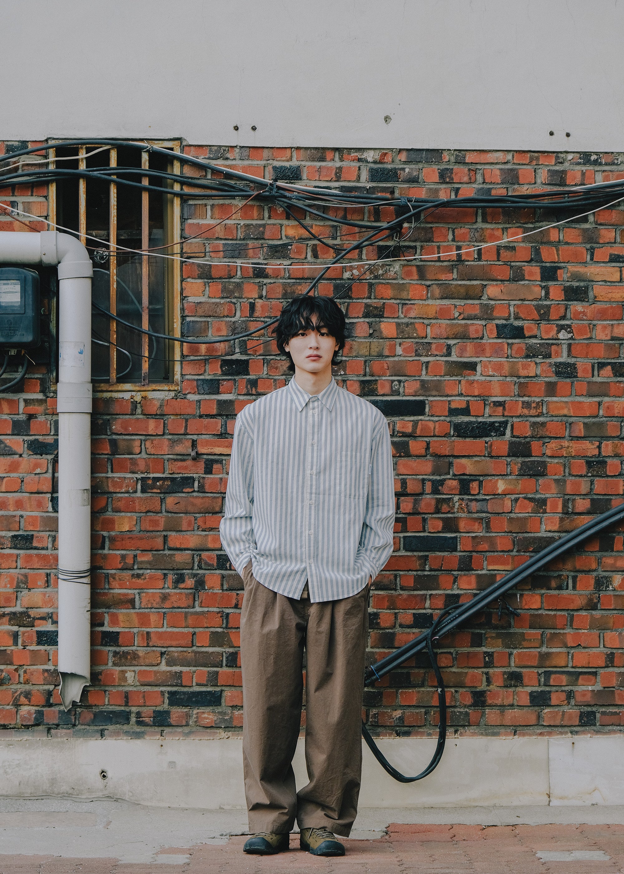 Person standing against a brick wall wearing a striped shirt and brown pants.