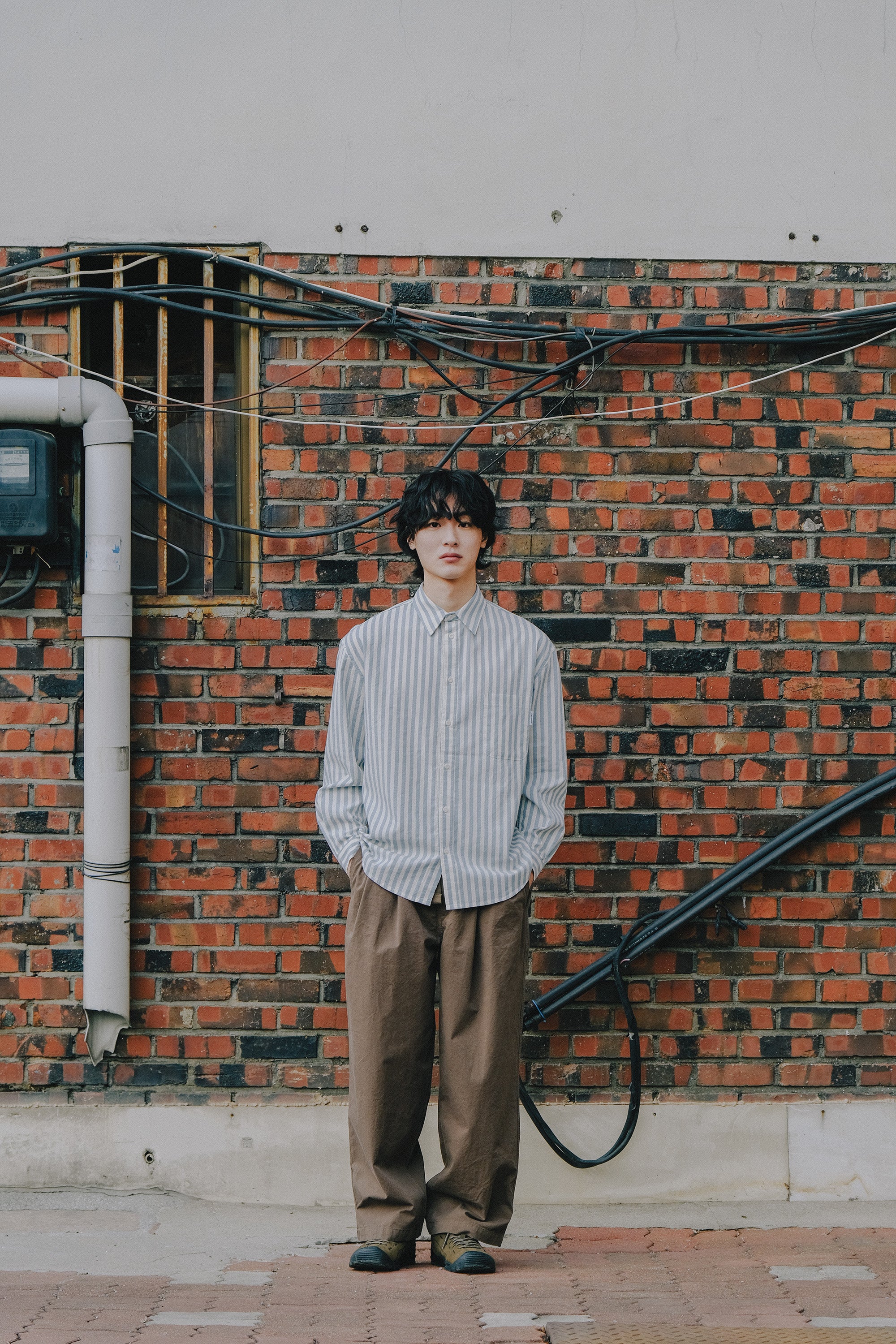 Person standing against a brick wall wearing a striped shirt and brown pants.