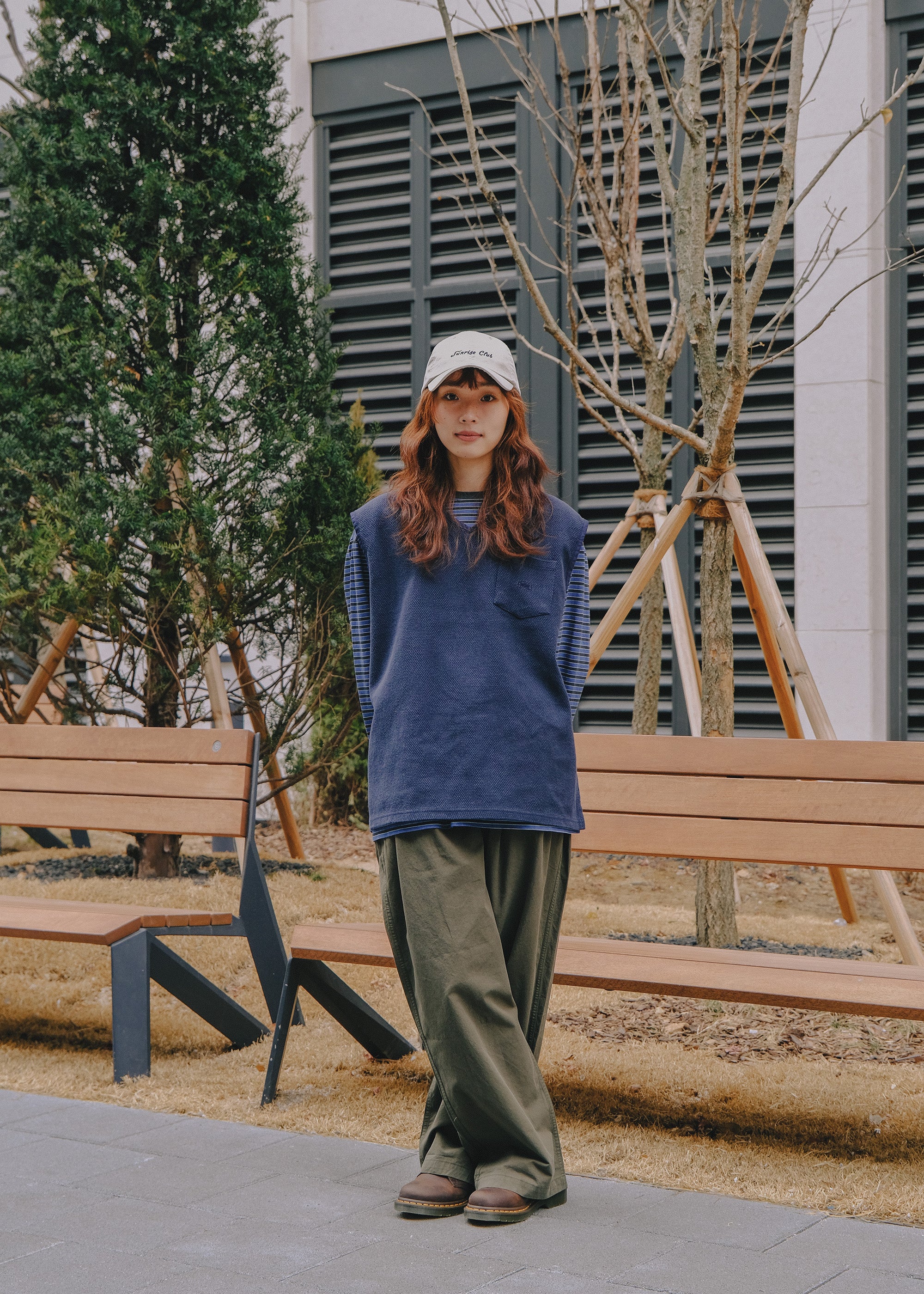 Person wearing a blue sweater and Brown pants standing outdoors near wooden benches and trees.
