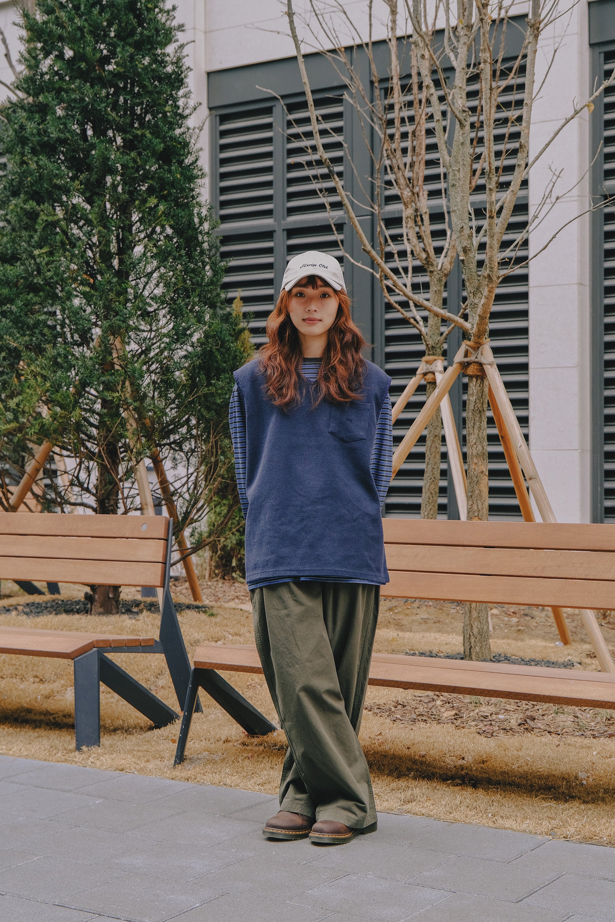 Person wearing a blue sweater and Brown pants standing outdoors near wooden benches and trees.