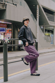 Person wearing a gray sweater and purple pants leaning against a railing in an urban setting.