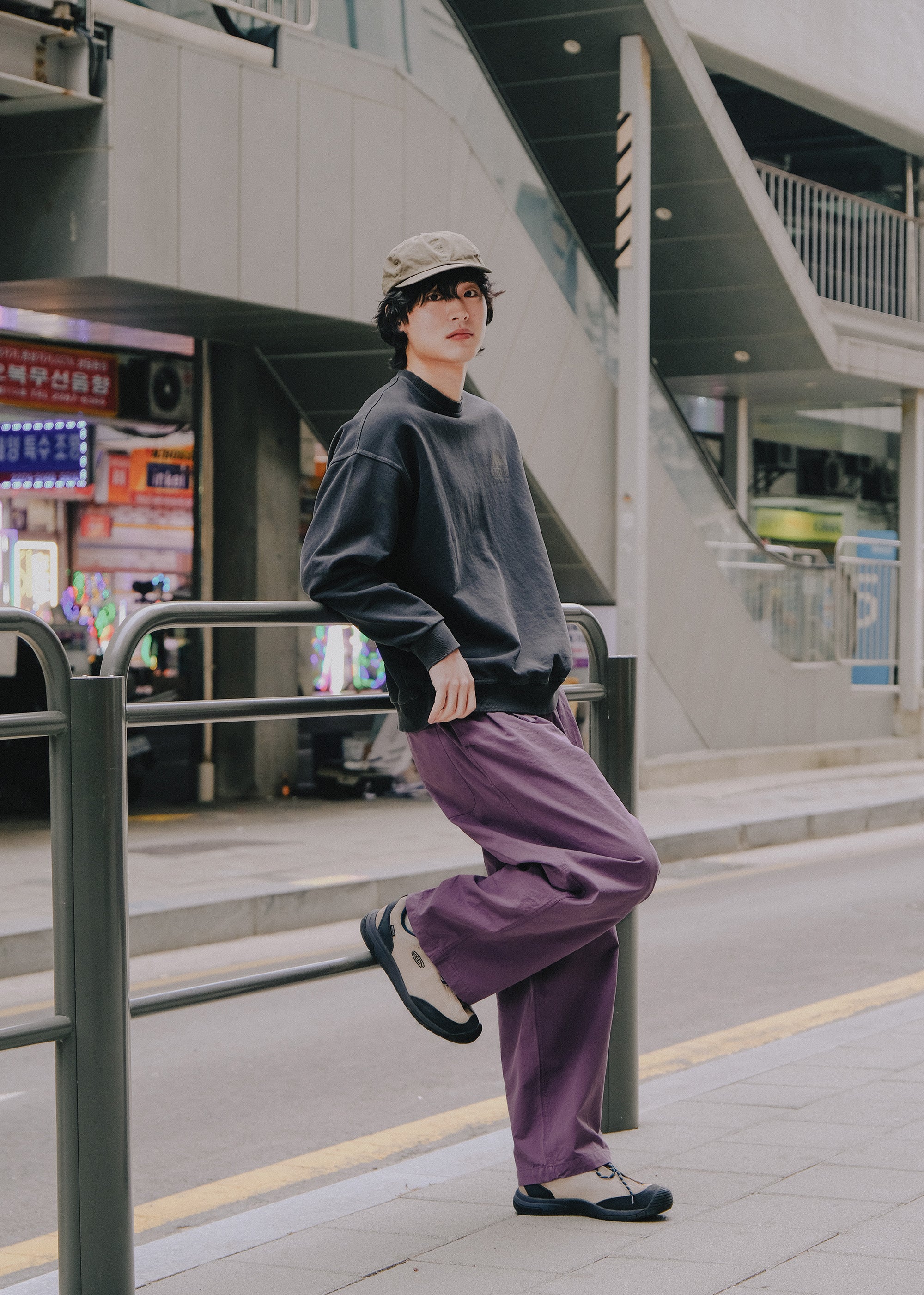Person wearing a gray sweater and purple pants leaning against a railing in an urban setting.