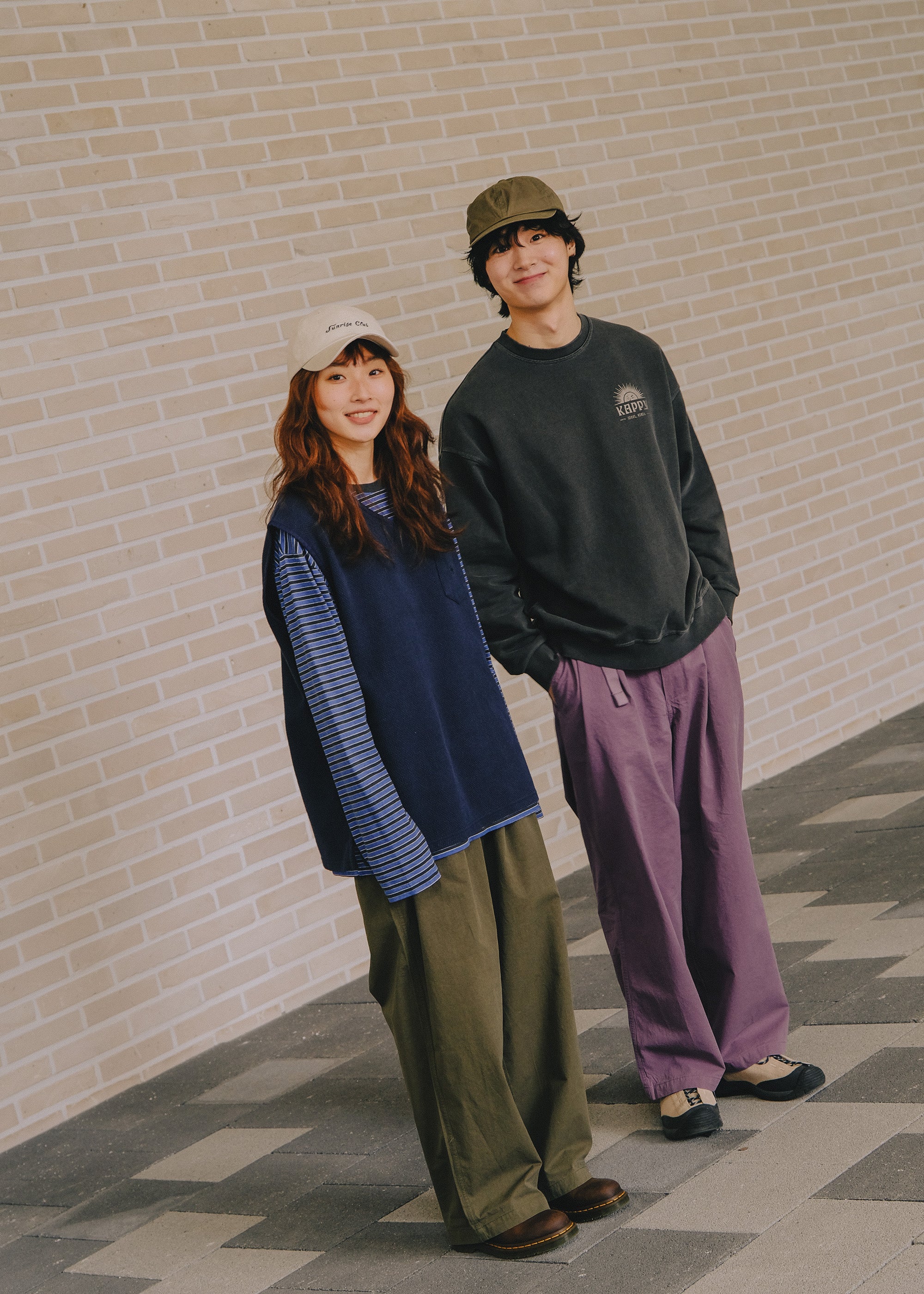 Two people standing against a brick wall, wearing casual clothing.