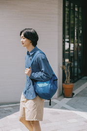 Man wearing a blue checkered shirt and beige shorts with a blue backpack, standing on a sidewalk.