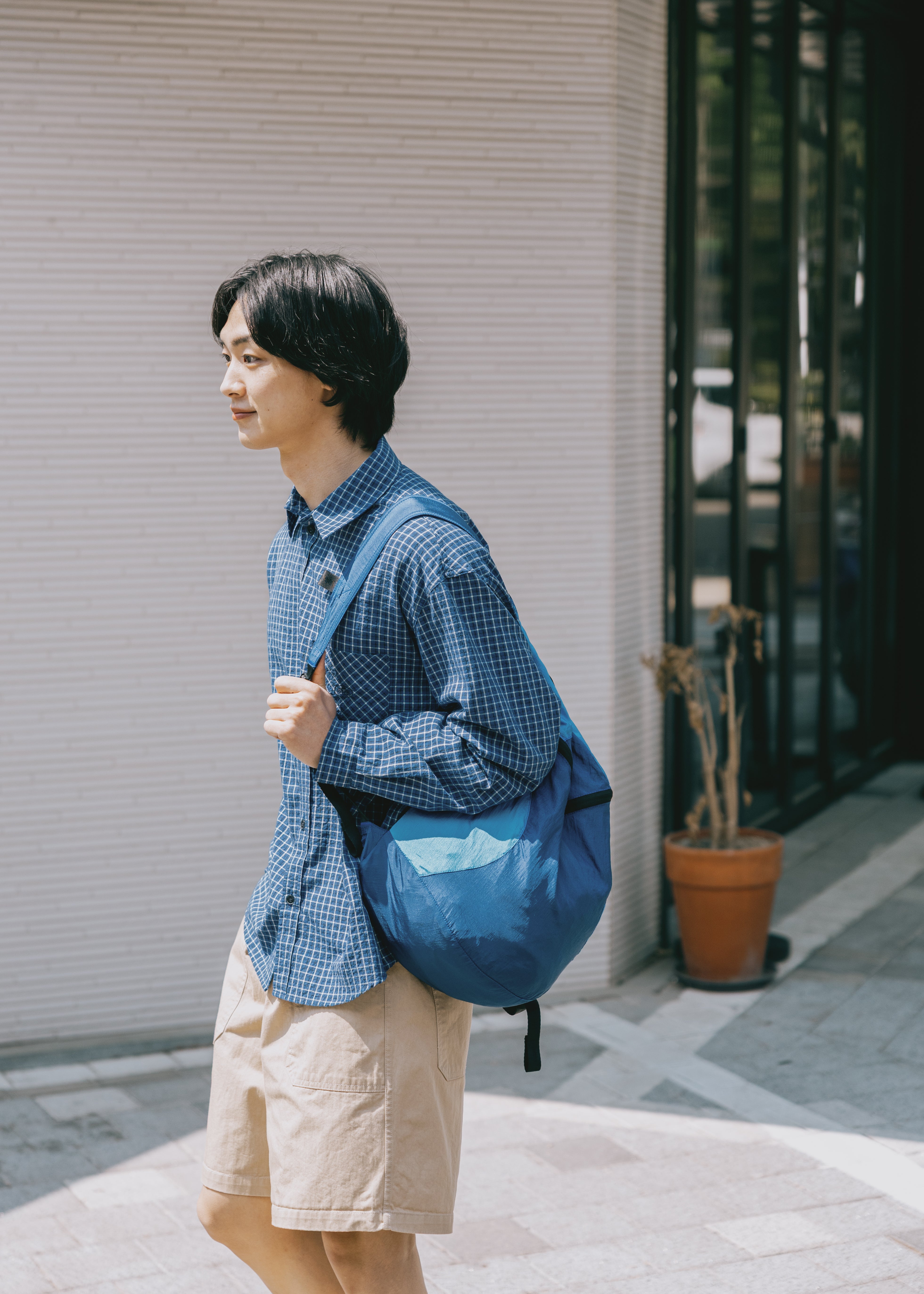 Man wearing a blue checkered shirt and beige shorts with a blue backpack, standing on a sidewalk.