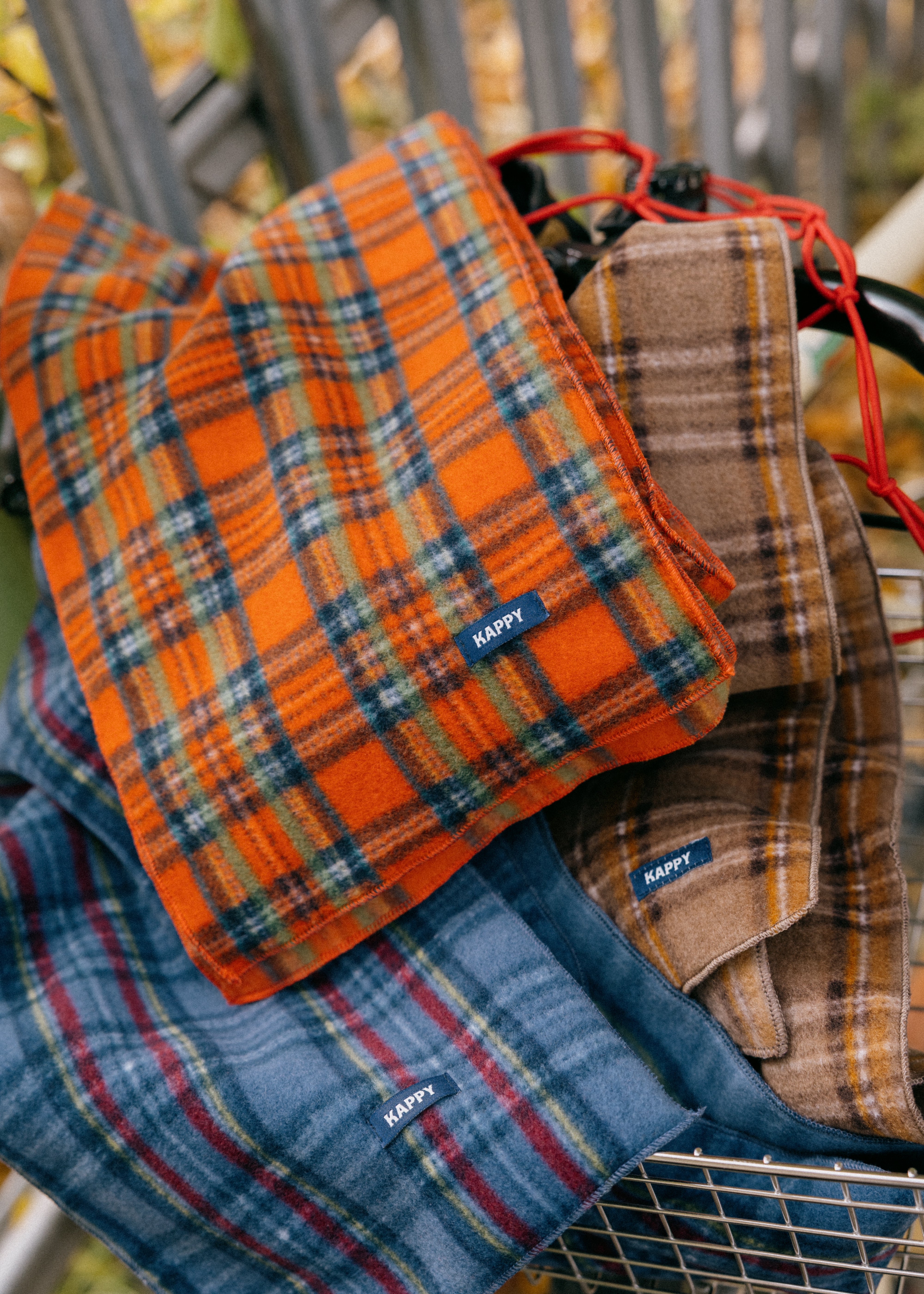 Three folded plaid fleece scarves  in orange, blue, and brown tones with  displayed in a wire basket outdoors. Each blanket has a small blue tag reading KAPPY.