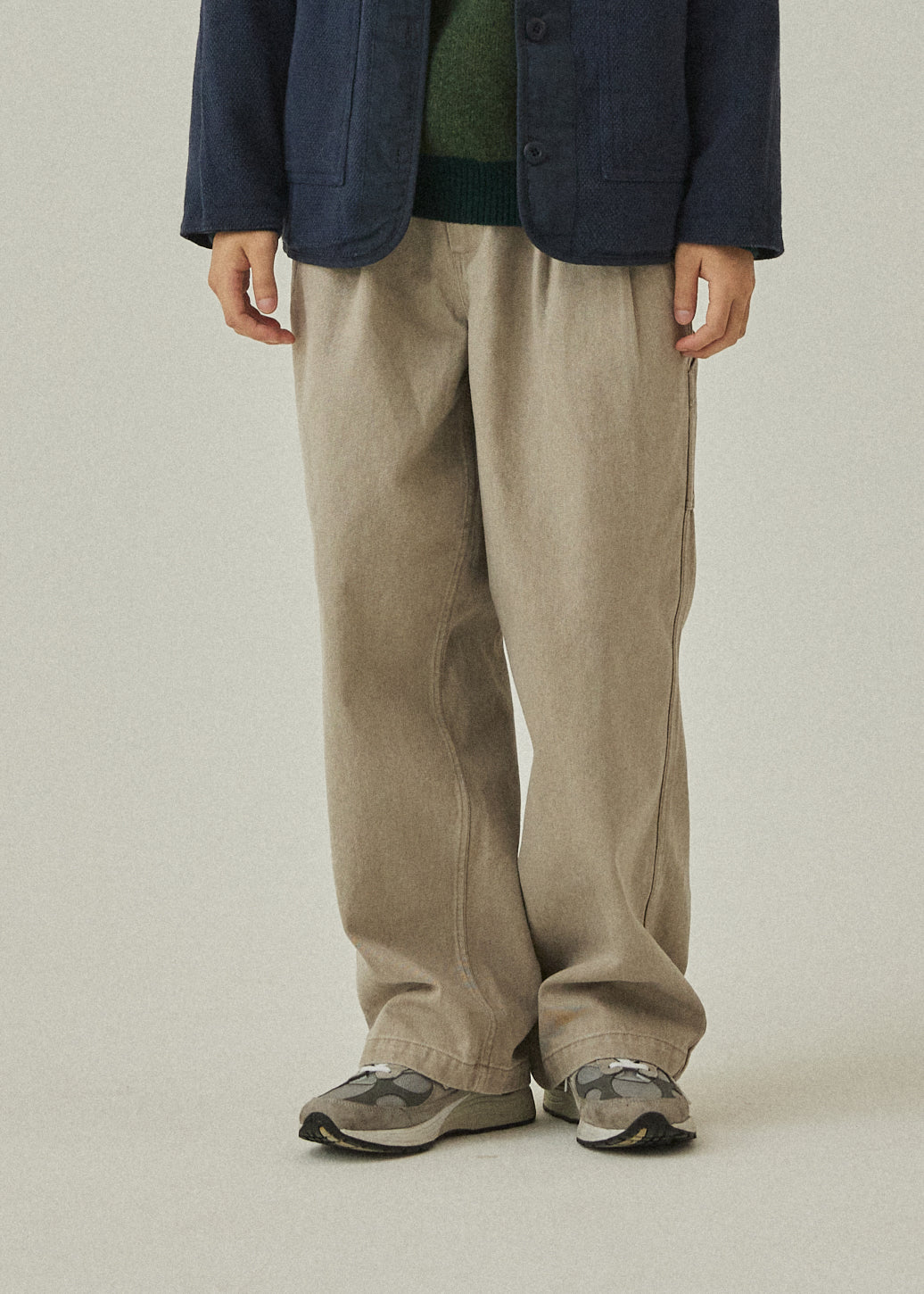 Person wearing a navy jacket, green shirt, beige pants, and gray shoes on a plain background