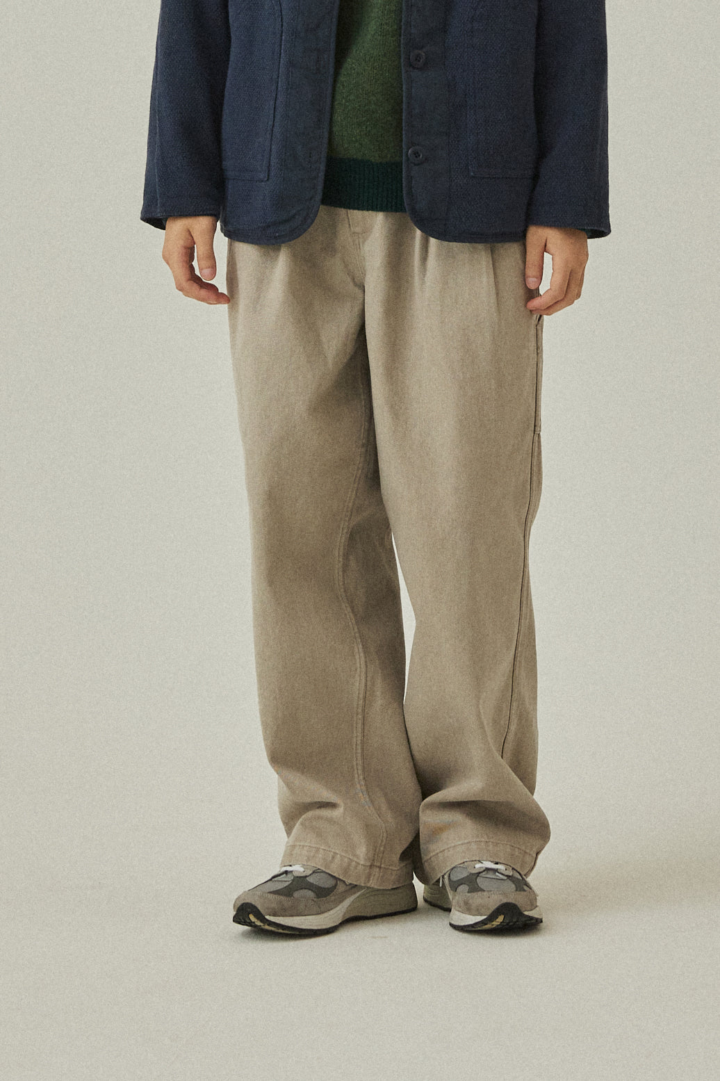 Person wearing a navy jacket, green shirt, beige pants, and gray shoes on a plain background