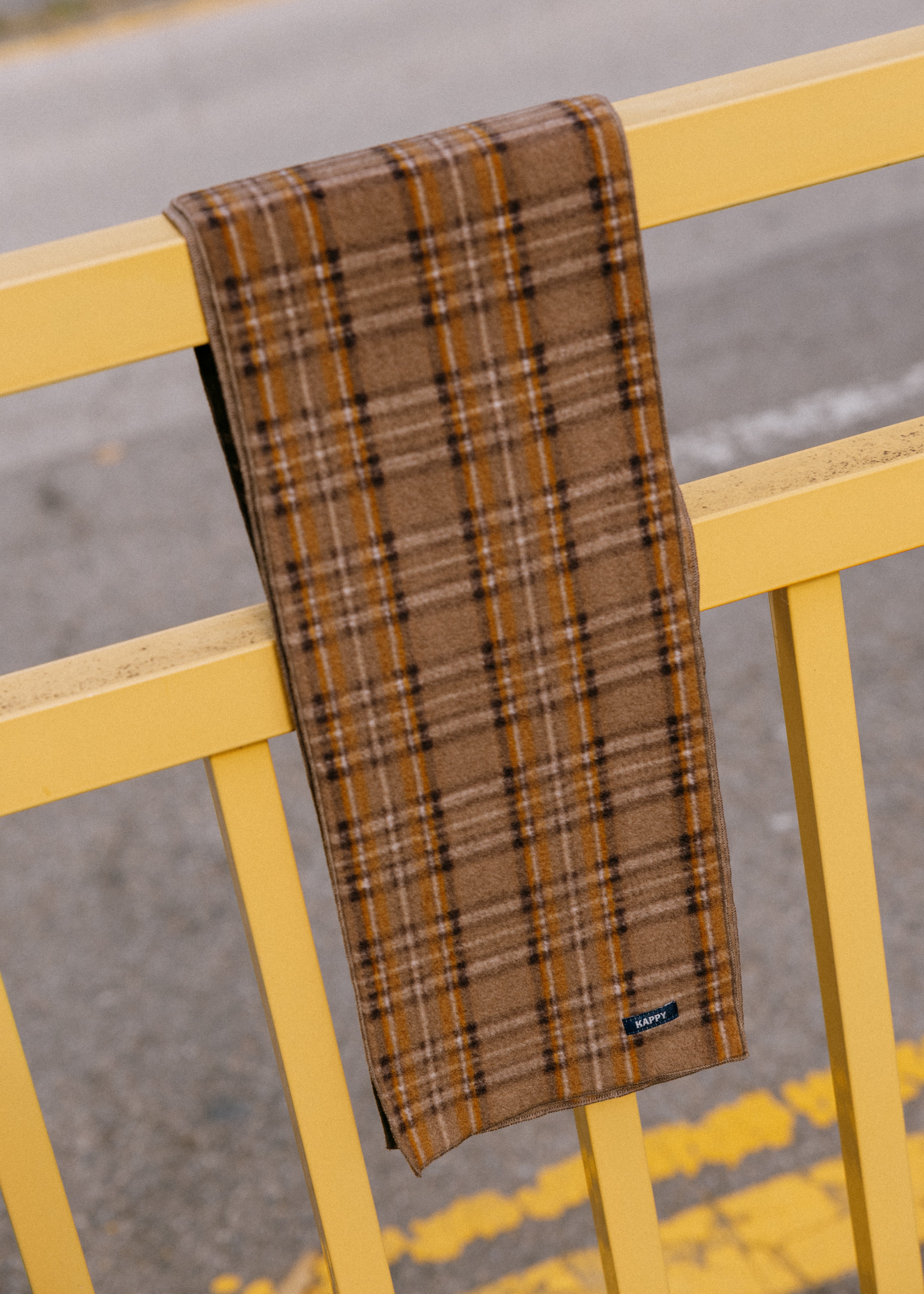 A brown plaid scarf draped over a yellow metal railing outdoors, with a gray asphalt road and yellow road markings in the background.