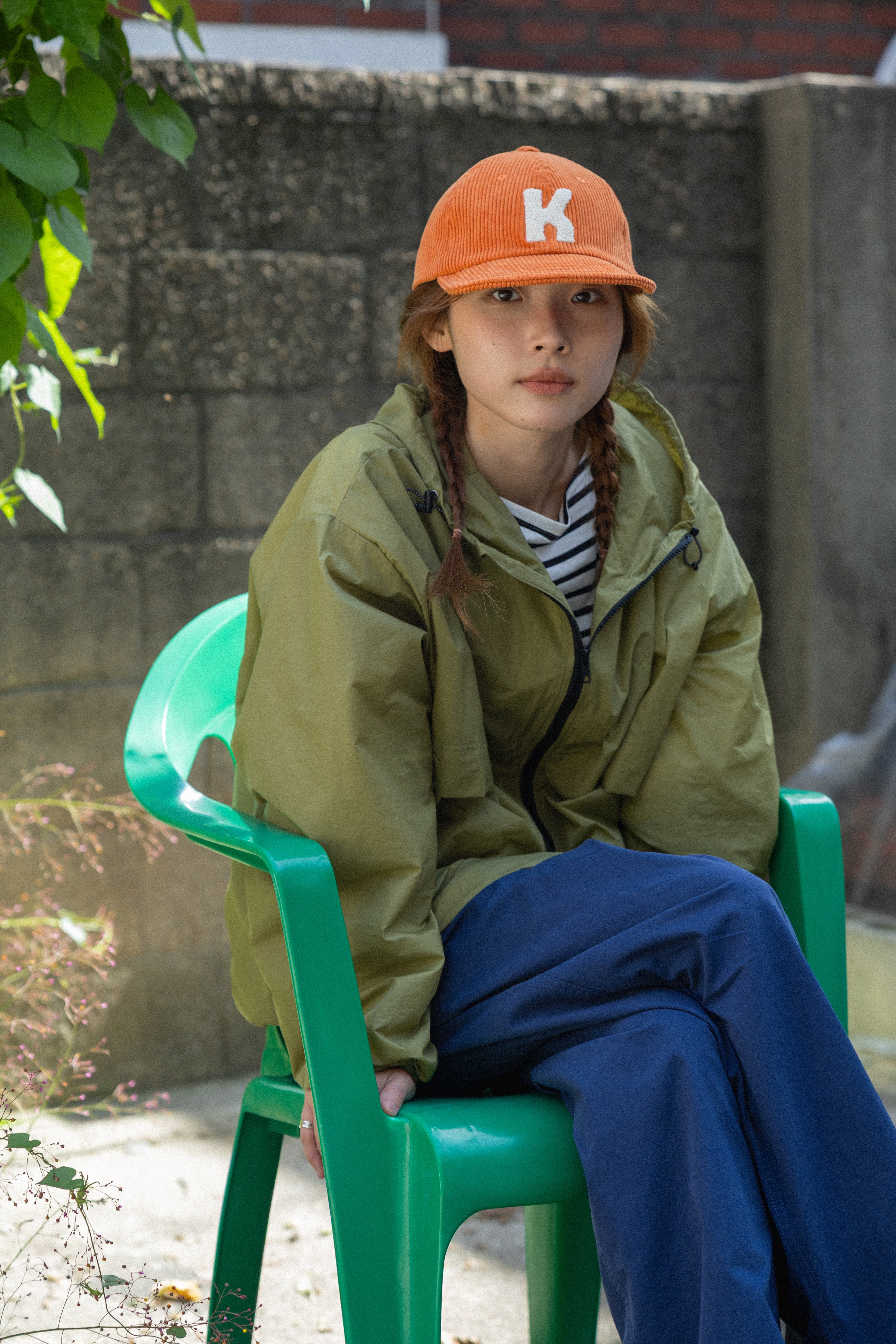A person sitting on a green plastic chair outdoors, wearing an olive green jacket, blue pants, a striped shirt, and an orange baseball cap with a white letter K.