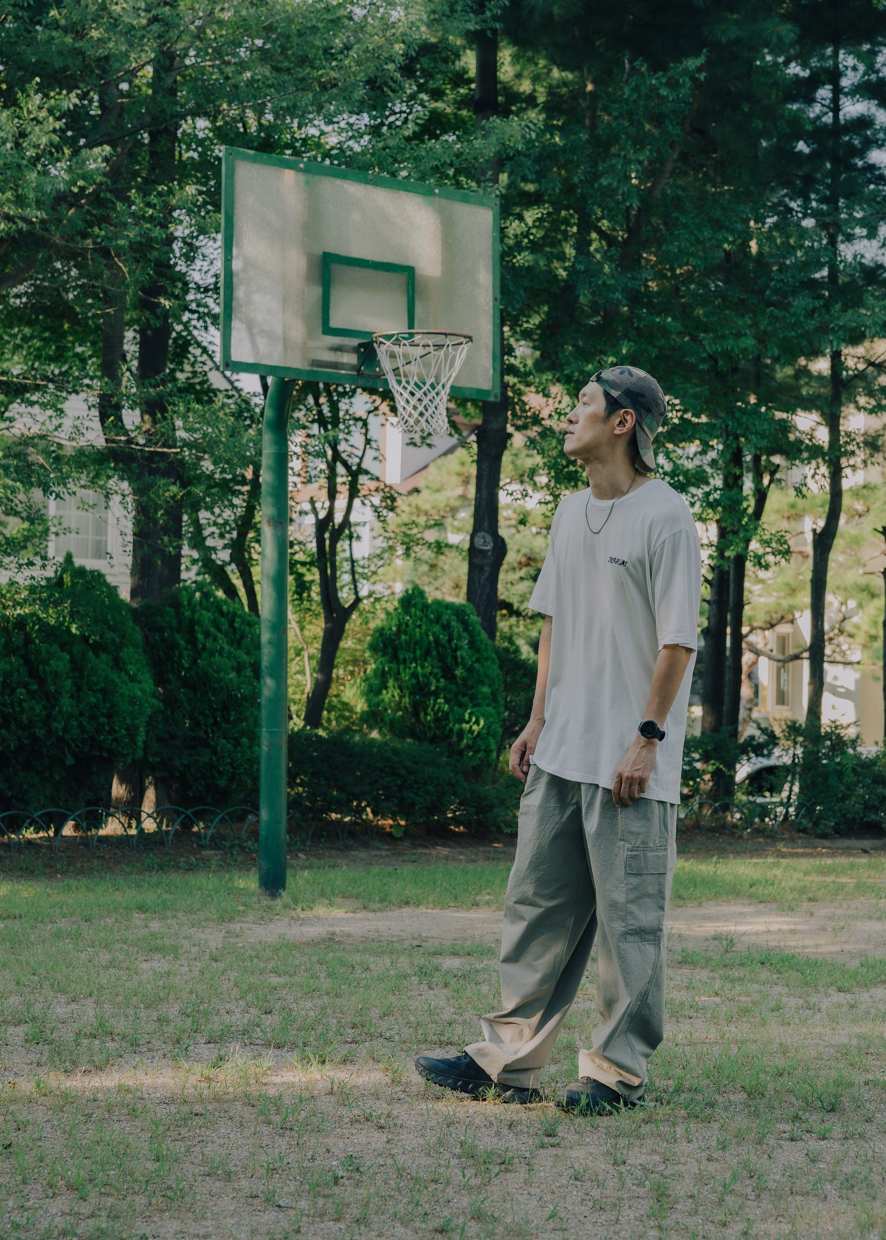 A man stands on a grassy outdoor basketball court looking up at a basketball hoop. He is wearing a light gray t-shirt, loose-fit khaki beige cargo pants, and a dark beanie.