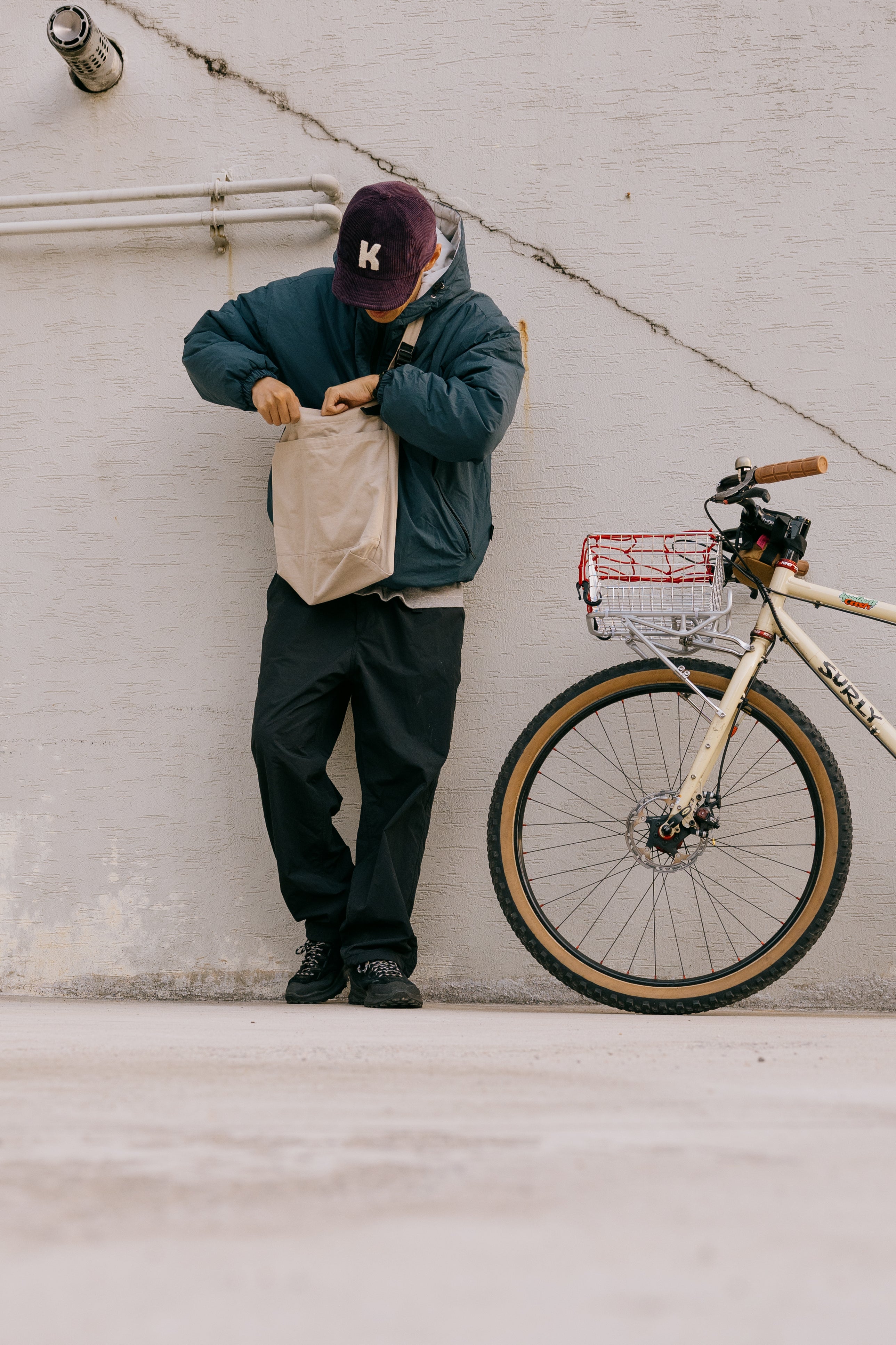 A person in a dark blue jacket, black pants, and a burgundy cap with a white 'K' stands against a concrete wall next to a cream-colored bicycle, holding a beige paper bag.