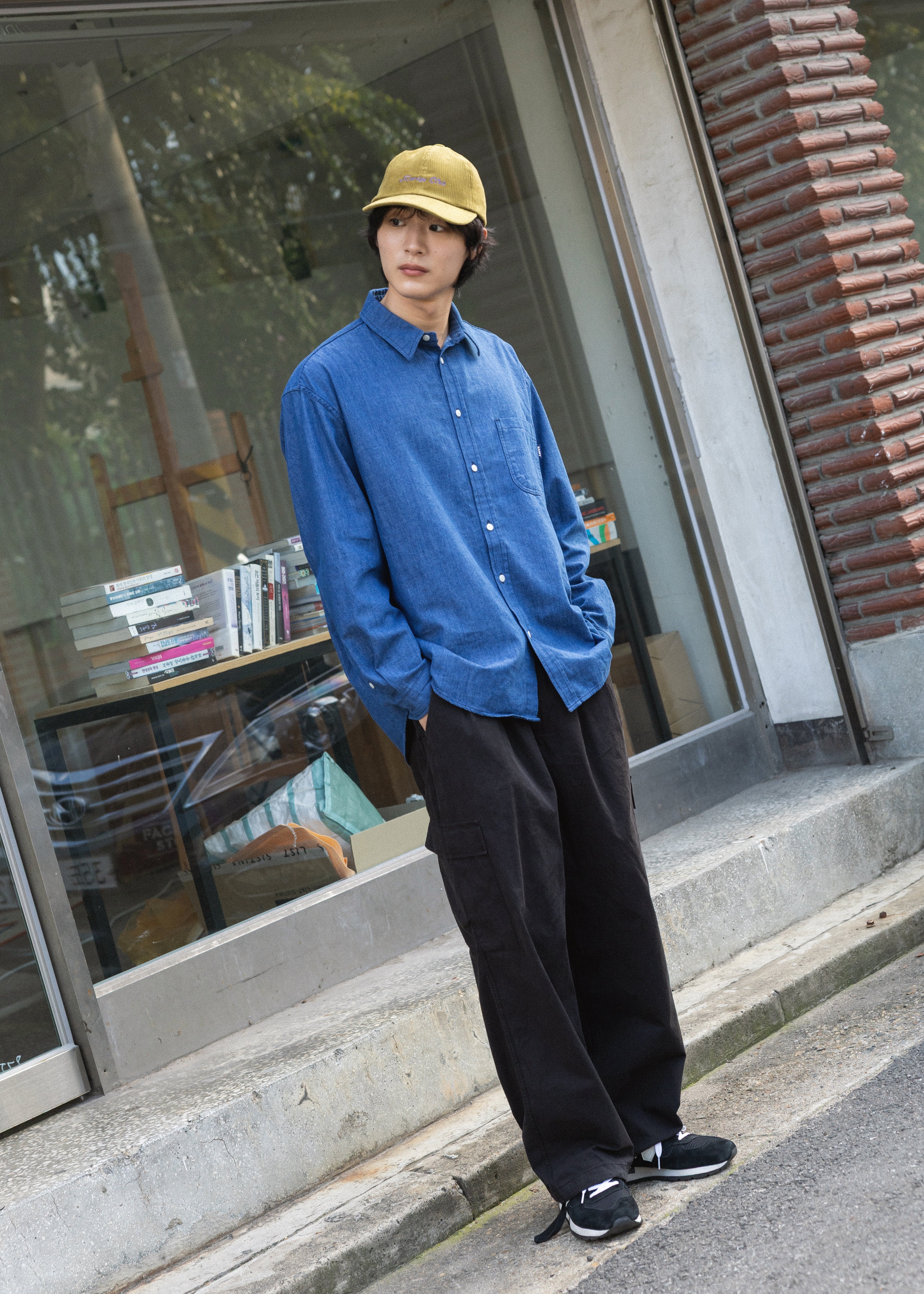A man wearing a blue long-sleeve button-up shirt, black wide-leg pants, black sneakers, and a yellow baseball cap stands on concrete steps outside a storefront with large windows and a brick column.
