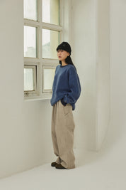 Person wearing a blue sweater and beige pants standing in a minimalistic room with a window.