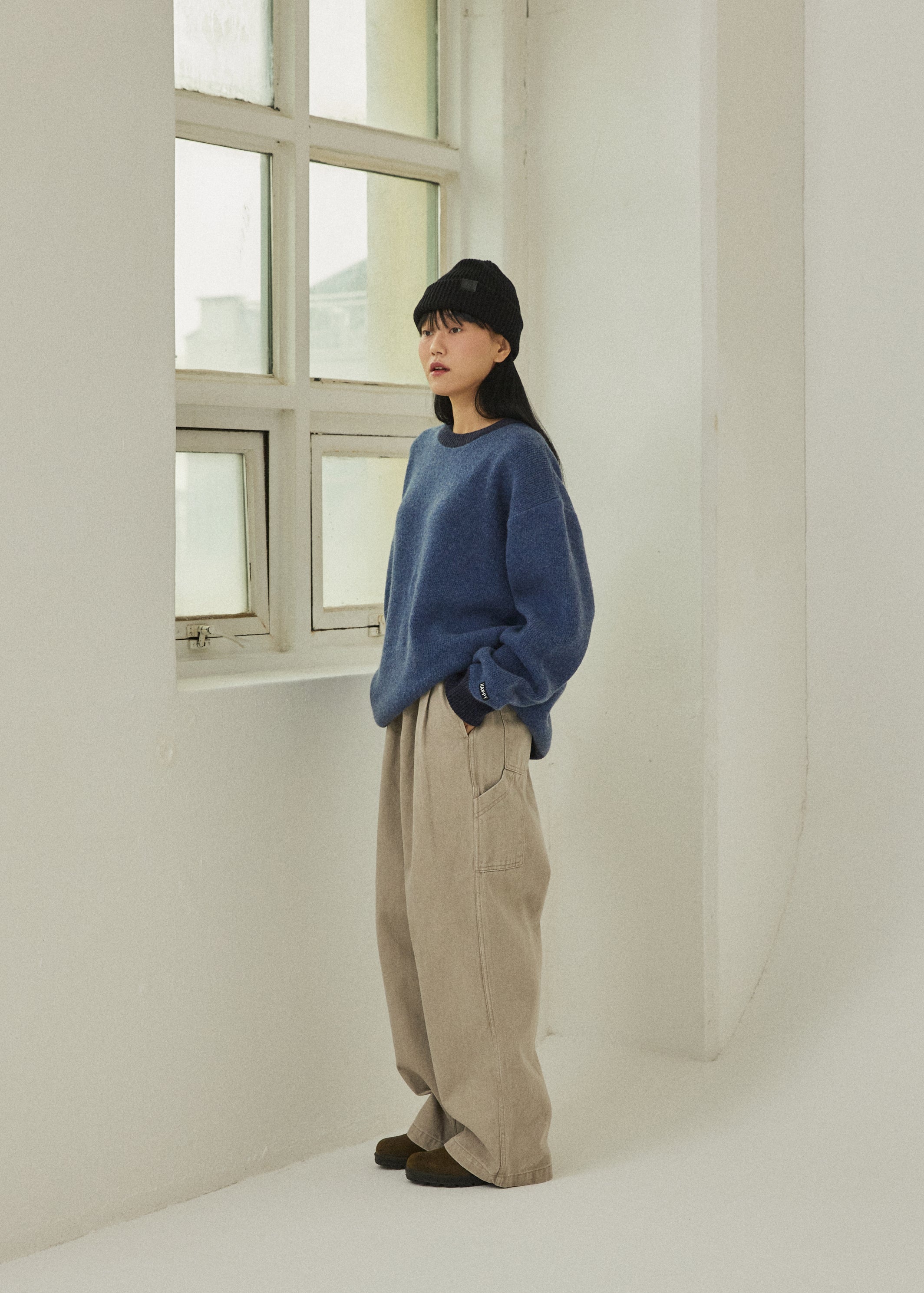 Person wearing a blue sweater and beige pants standing in a minimalistic room with a window.