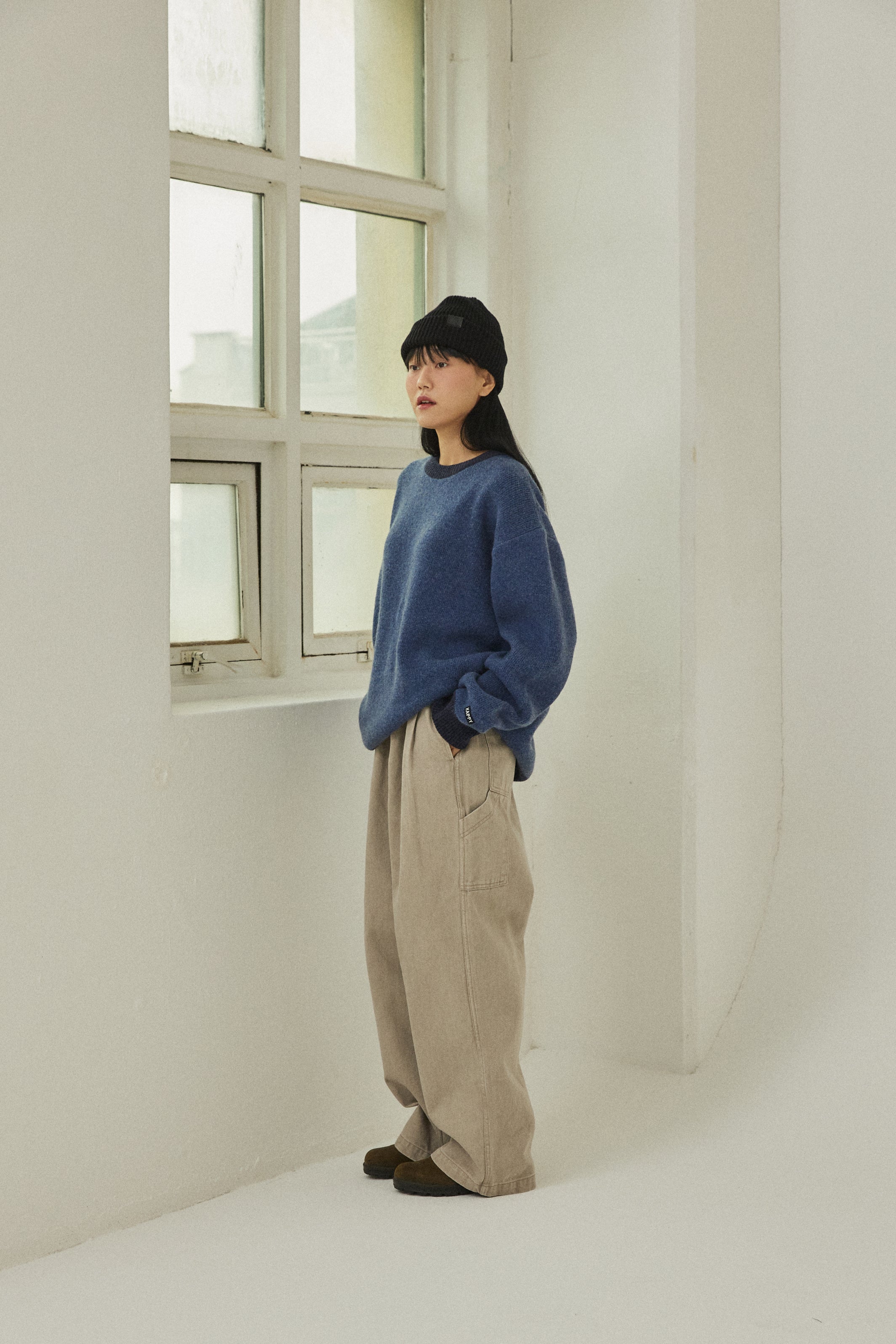 Person wearing a blue sweater and beige pants standing in a minimalistic room with a window.