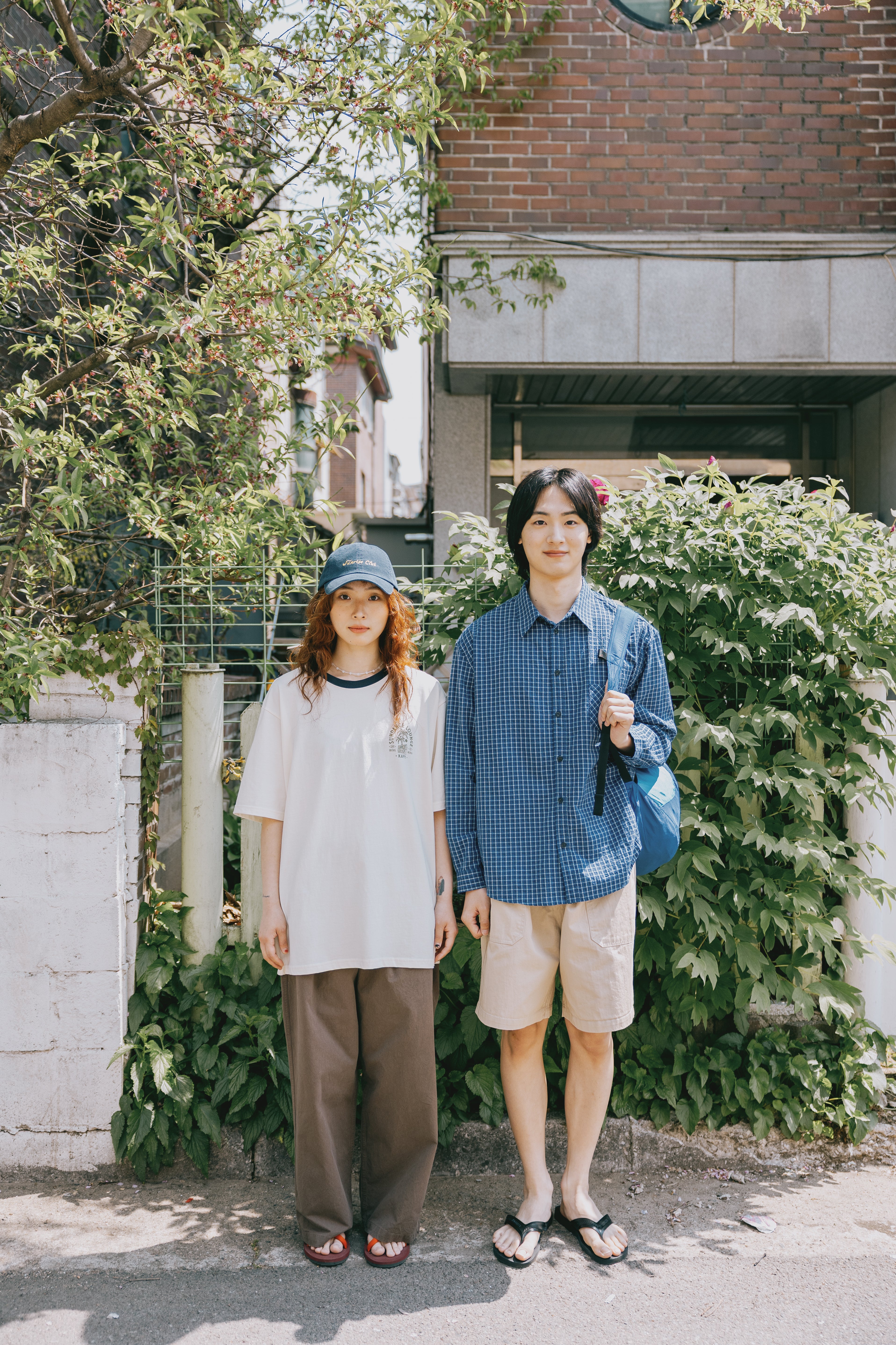 Two people standing outdoors with greenery and a building in the background